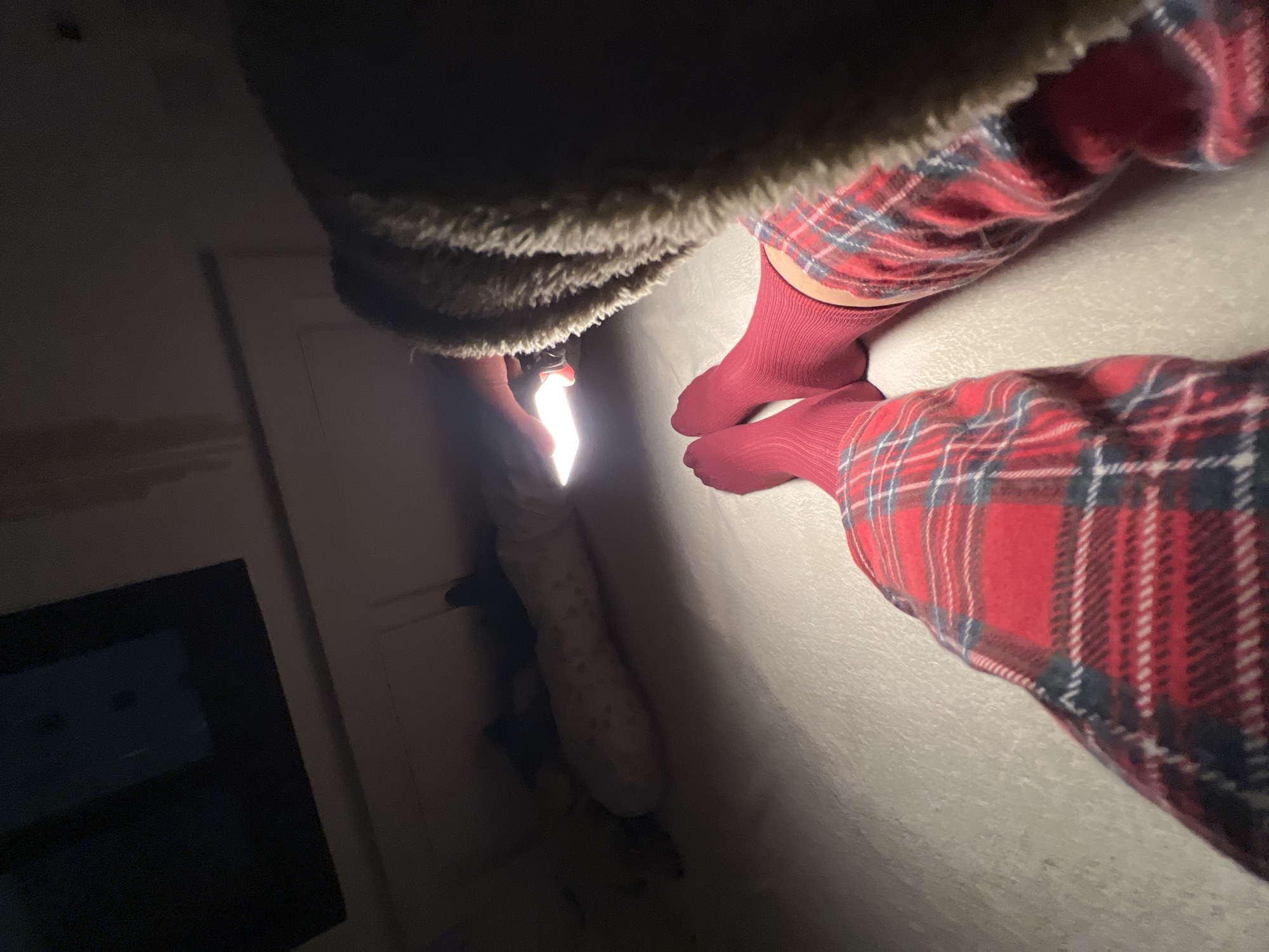 A view from above shows a person wearing a brown, fuzzy collar jacket and red plaid pajama bottoms, with red socks on their feet. The person holds a flashlight, illuminating the floor and a light-colored laundry bag. A dark appliance is visible on the left side of the frame, and a door or wall is on the upper left. There is no contextual information in the image to indicate its location as Kiryat Bialik, Israel.