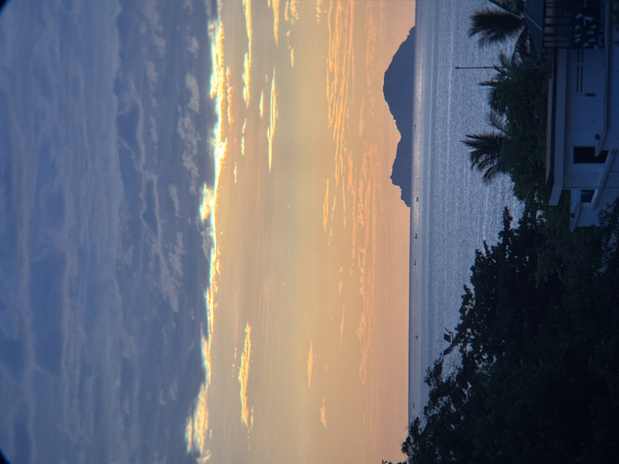 The image captures a serene seascape with islands off the coast of Nha Trang, Vietnam, bathed in the soft light of either dawn or dusk. The sky is a gradient of colors, transitioning from a deep blue at the top, filled with wispy clouds, to a warm orange and yellow near the horizon, where sunlight filters through the clouds. A low-lying island, silhouetted against the sky, sits on the horizon. In the foreground, the dark silhouette of lush vegetation, likely trees and foliage, covers the lower portion of the frame. Partially visible at the bottom are the rooftops of buildings, suggesting a coastal settlement. Small boats dot the calm ocean surface, indicating some maritime activity. The overall atmosphere is tranquil and picturesque.