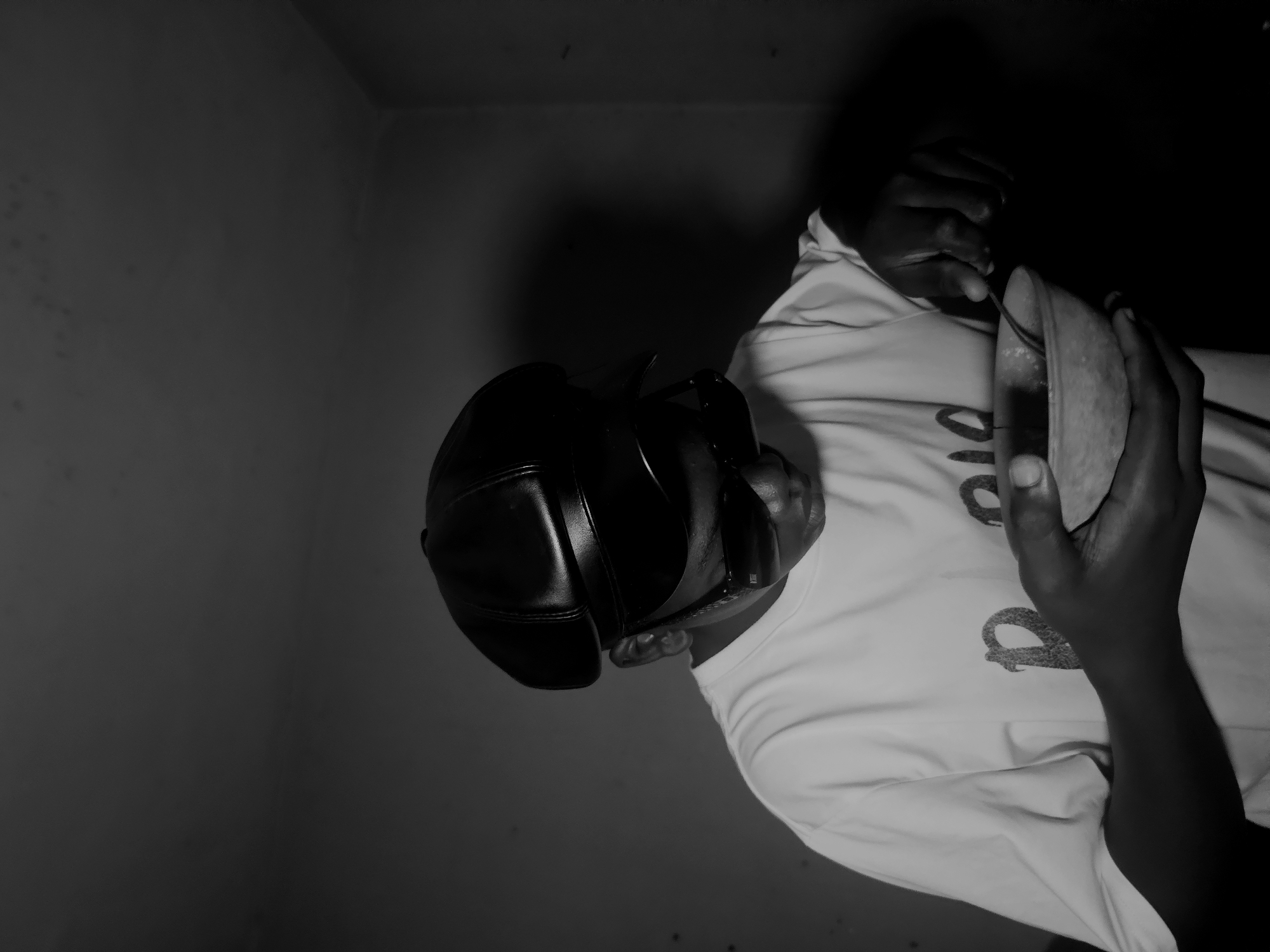 A black and white photograph depicts a person, presumably in Nkwelle, Nigeria, wearing a baseball cap and a white t-shirt with a graphic on it. The person is holding a halved pear in their hands, appearing to be examining or eating it. The background shows walls, suggesting an indoor location within a building. The lighting is dim, creating strong contrasts between light and shadow. The photograph is rotated, with the subject oriented sideways.