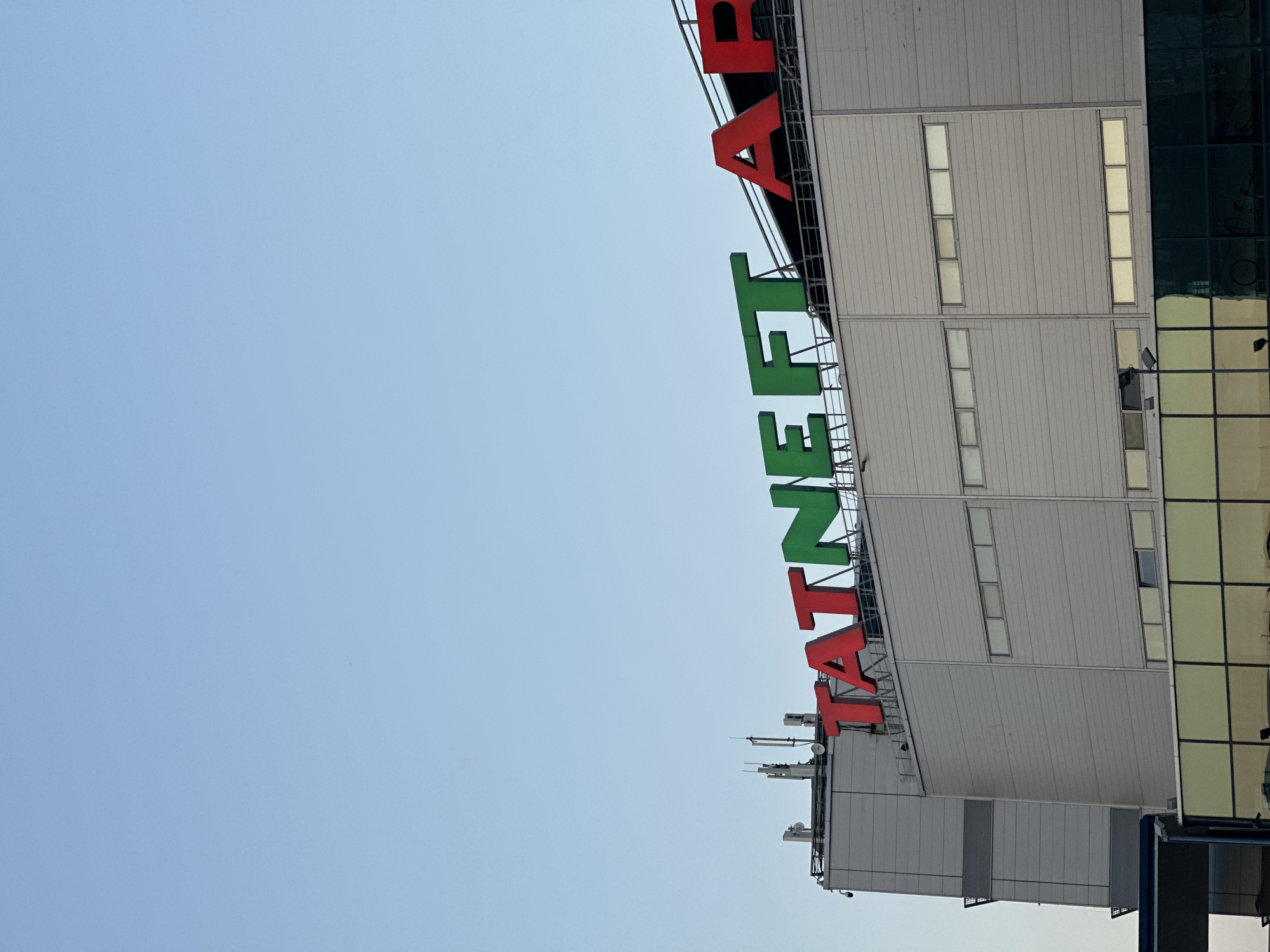 The image is of the "Tatneft Arena" building in Kazan, Russia. The building has a large sign that says "Tatneft Arena" in big red and green letters. The building is gray with several rows of windows. There is also some antenna on top of the building. The sky above is light blue.