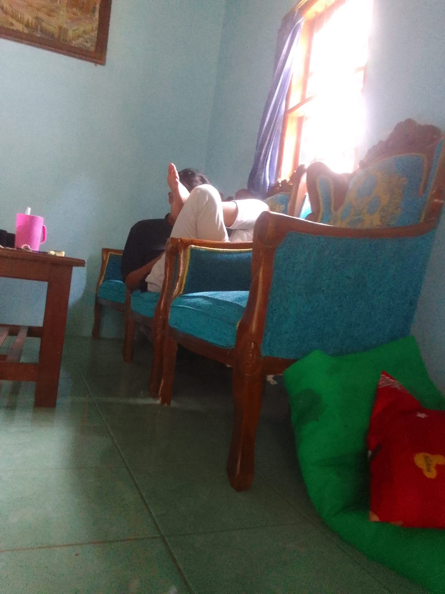 In a dimly lit room with pale blue walls, a person is seated on a turquoise armchair. The person is wearing white pants and a dark shirt. Their legs are propped up, and their feet are visible. The armchair has wooden trim. To the left of the armchair is a small wooden table with a pink cup on it. In the background, a window is illuminated by bright sunlight, with a purple curtain drawn to the side. A large green pillow with a red pillow on top is situated on the floor in front of another armchair. The floor appears to be tiled. The scene is located in the city of Suren, Indonesia.