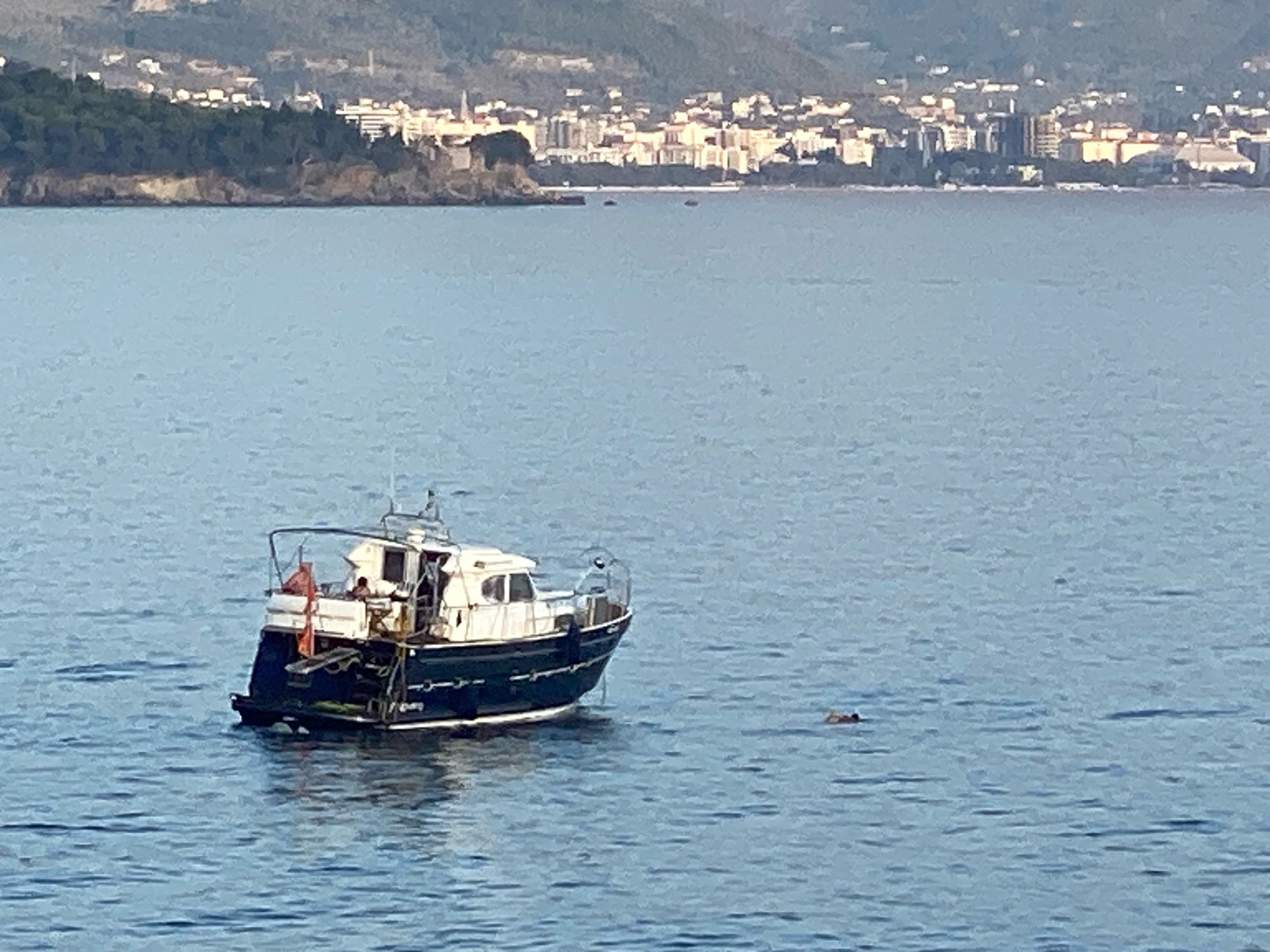 In the calm, blue waters of the Adriatic Sea, a dark blue and white motorboat is anchored. A person is visible on the deck of the boat. To the right of the boat, a person is swimming in the sea. In the background, the coastal city of Sutomore, Montenegro, is visible, with buildings and green hills lining the shoreline. The sky is clear and the sun is shining.