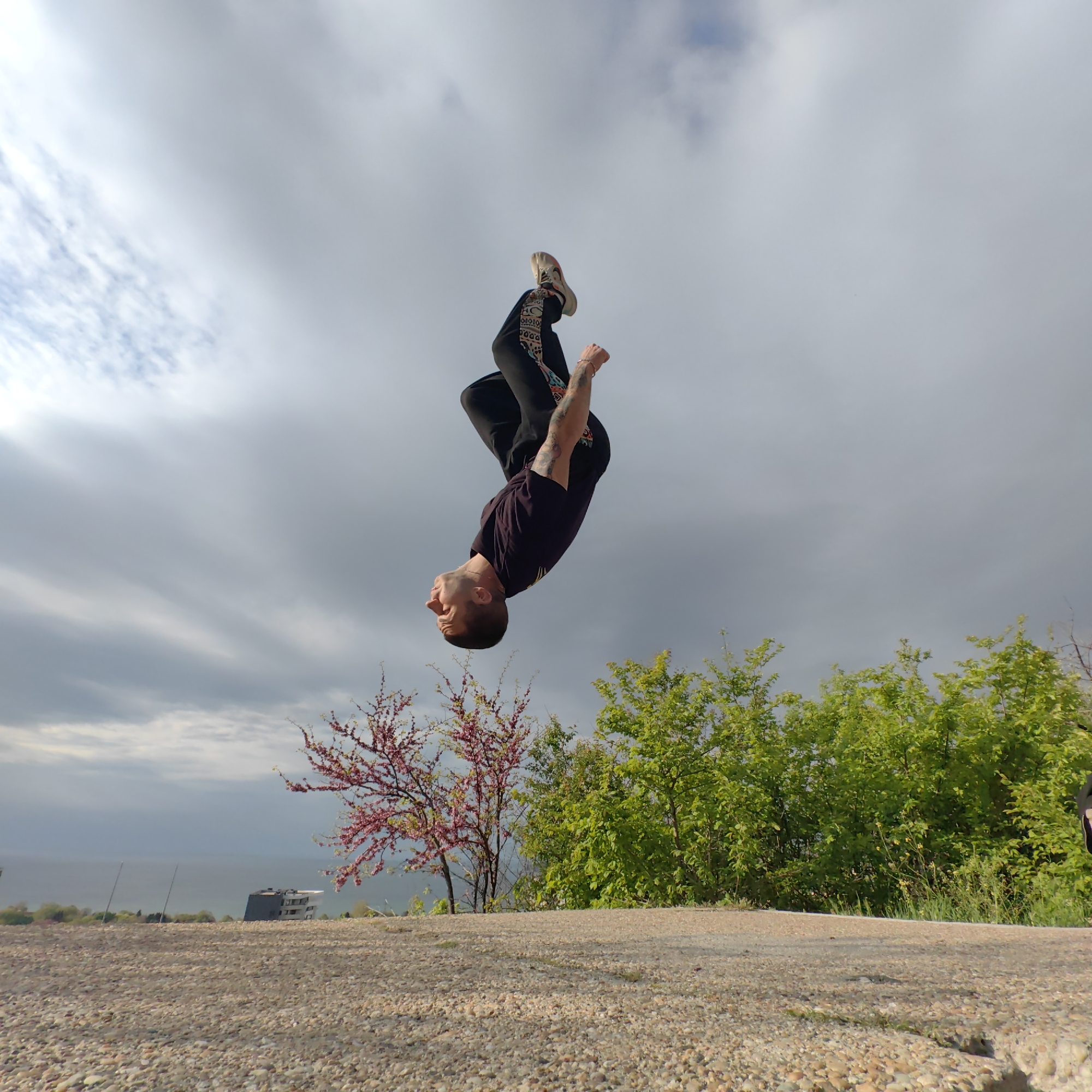 A man is performing a backflip in a park in Varna, Bulgaria. The background features a cloudy sky, trees, and a distant view of the sea. The foreground consists of a gravel or concrete surface. The man is wearing dark pants with patterned details, a dark t-shirt, and light-colored sneakers. He has tattoos on his arm. The setting is likely a recreational area with natural elements combined with urban infrastructure. A building is visible near the horizon and a tree with pink blooms is located on the left, while green leafy trees are located to the right.