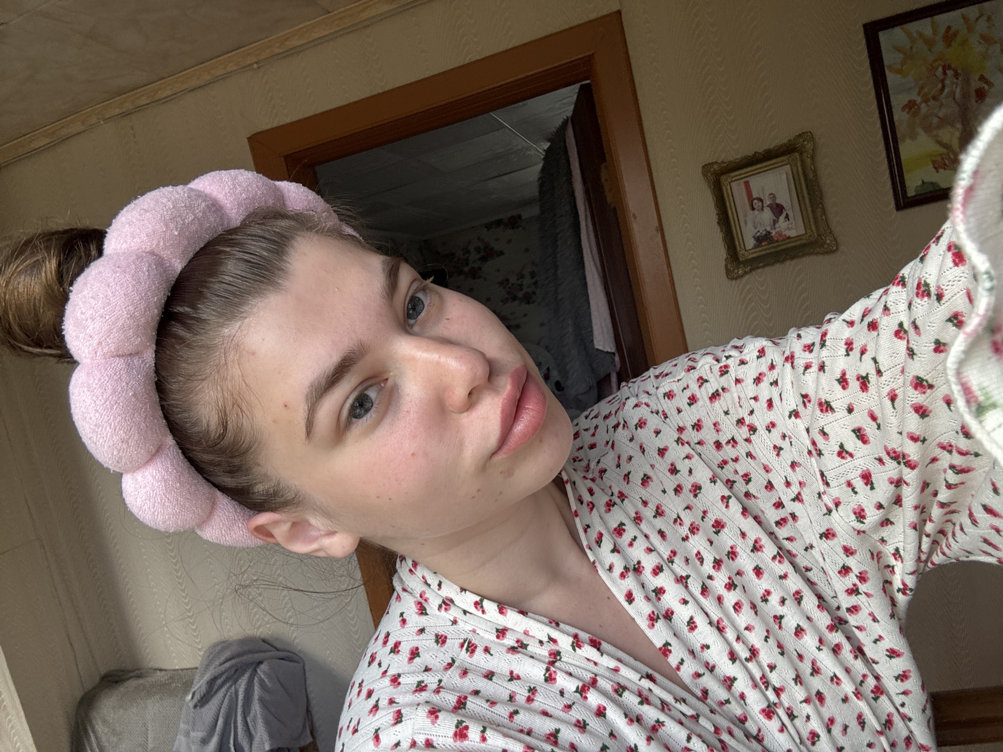 A young woman with fair skin and light eyes is wearing a pink padded headband and a white floral-patterned robe or top. Her hair is pulled back into a bun. She is positioned indoors, likely in a home, as suggested by the wallpaper, framed pictures on the wall, and a doorframe in the background leading to another room with floral wallpaper. The location cannot be determined from the image.