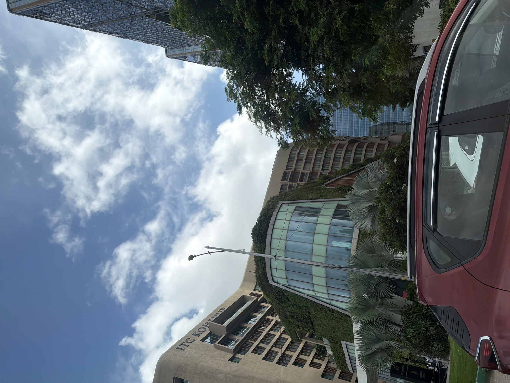 The image depicts a modern architectural complex in Hyderabad, India, featuring buildings with contemporary designs and extensive greenery. The prominent structure on the left appears to be a hotel or corporate building, identified by the signage "ITC KOPARAL" visible on its facade. This building is characterized by its curved shape and is extensively covered in vertical gardens, with lush green foliage cascading down its exterior. A section of the building showcases a striking glass-curved facade, reflecting the surrounding environment and sky.
To the right of this main structure, another tall, modern building with a glass facade and geometric patterns is visible, suggesting a commercial or office space. The foreground is partially occupied by the roof and rear section of a dark red car, with its windows tinted. Behind the car, ornamental palm trees and other dense greenery create a tropical ambiance. The sky above is a vibrant blue, dotted with fluffy white clouds, indicating a daytime scene. The overall impression is one of a well-designed urban landscape that integrates nature with modern architecture.