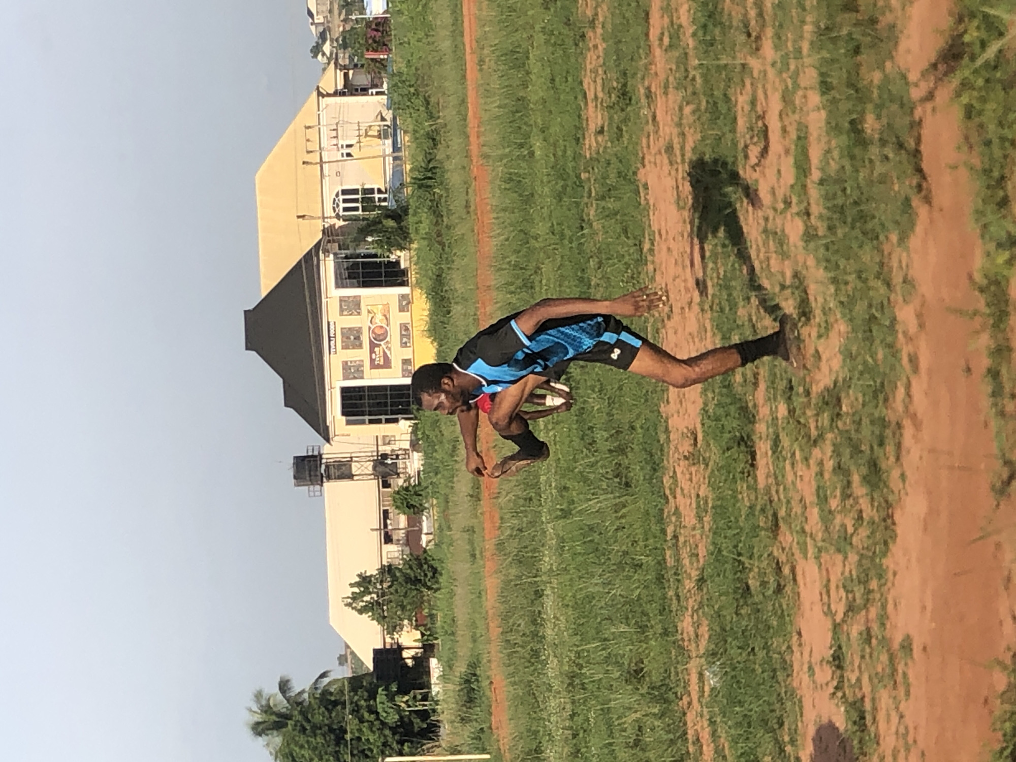 The image displays an outdoor scene in Ukwu-Abwa, Nigeria, featuring a male individual in an athletic stance on a field under bright daylight. The man, with dark skin and short dark hair, is wearing a black and blue short-sleeved athletic top with blue patterns and matching black shorts. He is captured in a dynamic, crouched pose, leaning forward with bent arms and spread legs, suggesting he is in the middle of a jump, a sprint start, or a landing.
The setting is an open field with varying textures of green grass and reddish-brown dirt. A clear, long shadow of the man is visible on the ground behind him, indicative of direct sunlight, likely during the morning or late afternoon. The sky above is clear and a pale grey-blue.
In the background, several structures are visible. A prominent building features a light yellow or beige facade, multiple windows, and a distinctive dark roofline. The lower section of this building has what appears to be a large mural or sign, on which some text, possibly "KEVOLI FOODS," and other graphic elements can be faintly discerned. Other buildings, some with water tanks on their roofs, and scattered trees and vegetation complete the background, creating the impression of an urban or semi-urban environment bordering an open space. The overall atmosphere is bright and active.