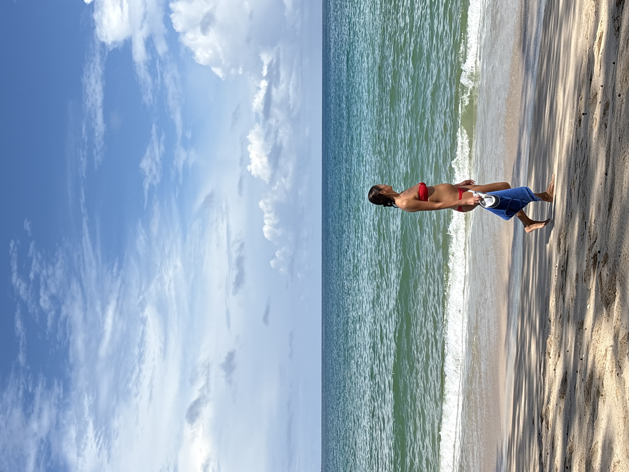 A woman in a red bikini walks along the sandy shore of a beach in Choeng Thale, Thailand, beside the turquoise ocean. She is carrying a white cylindrical object and a folded blue towel in her left hand. The sky above is a bright blue with scattered white clouds. The sun casts shadows of trees onto the sand in the foreground.