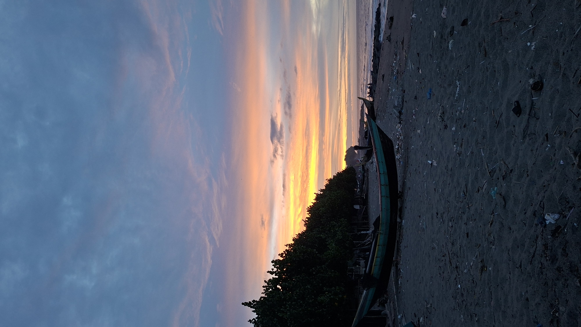 This is a landscape photograph taken at a beach in Lhokseumawe, Indonesia. The image is oriented vertically, with the sky taking up a significant portion of the left side and the beach occupying the right.
The sky is a dramatic canvas of a sunset, displaying vibrant streaks of orange and yellow blending into softer pinks and blues. Clouds are scattered across the sky, some wispy and others denser, catching the fading light. The sun appears to be low on the horizon, just above the tree line.
In the foreground, a sandy beach is visible, littered with debris, including plastic waste and natural detritus like twigs and leaves. To the right of the image, a long, narrow boat, painted green and red, rests on the sand, angled towards the water. Behind the boat, a dense line of dark green trees forms a lush border along the edge of the beach.
The scene suggests a quiet, possibly end-of-day setting. The presence of the boat and the shoreline indicates a coastal location. The debris on the beach hints at environmental concerns or the aftermath of human activity. The overall mood is serene, with the breathtaking sunset dominating the visual experience. There is no visible text in the image.