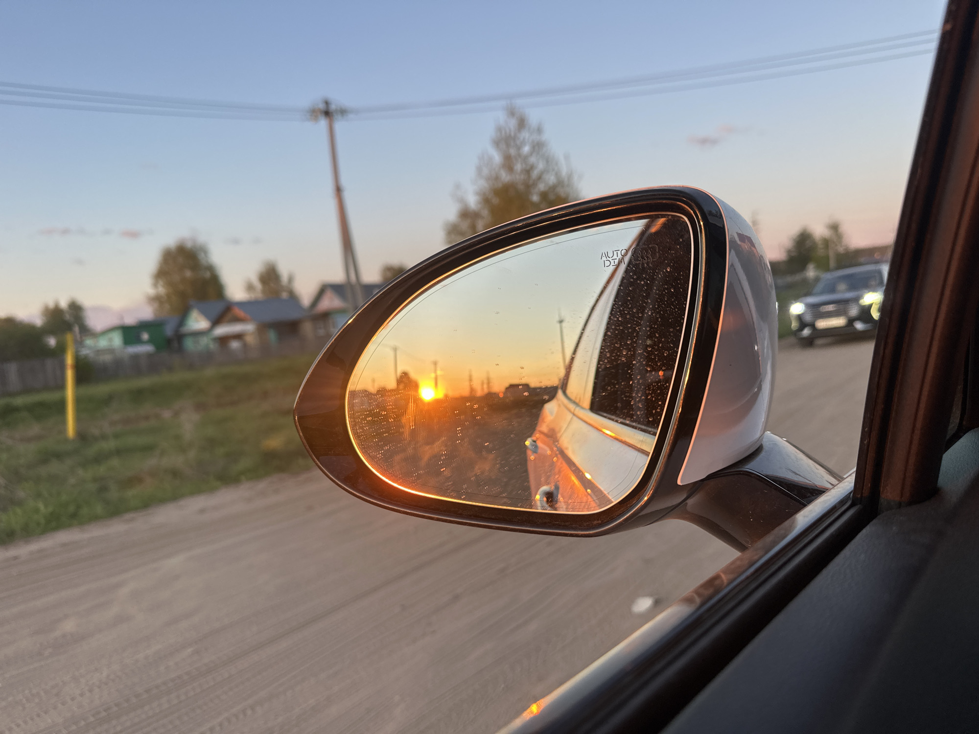 Captured from within a vehicle, the image foreground features a passenger-side rearview mirror reflecting a sunset. The sunset dominates the mirror's reflection with a bright, orange sun appearing on the horizon. In the background visible through the windshield, a dirt road stretches into the distance alongside a row of small, rural houses. A utility pole is seen alongside the road, near the houses. An SUV, likely an Audi, is positioned behind and to the right of the vehicle, its headlights illuminated. The location appears to be Chaikovsky, Russia, based on the architecture and general environment.