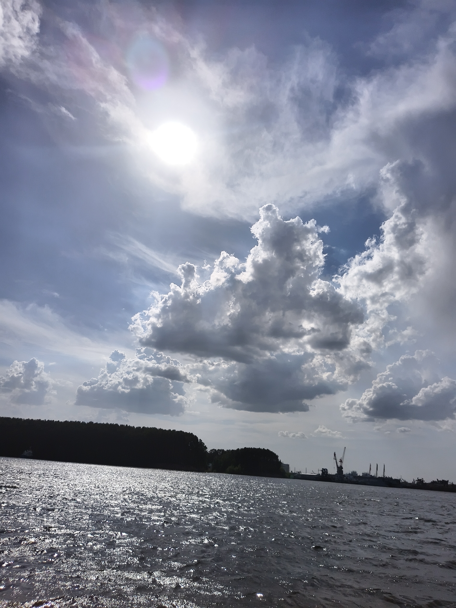 The image depicts a body of water, possibly a wide river, with sunlight reflecting off its surface, creating a shimmering effect. The water is dark. On the horizon, there is a dark silhouette of trees and low-lying vegetation. Further to the right, there appear to be industrial structures or machinery, including cranes and what appear to be docking facilities. Above, the sky is filled with dynamic cloud formations, ranging from light, wispy cirrus clouds to larger, more prominent cumulus clouds. The sun is visible, partially obscured by the clouds, with a bright halo. The image appears to have been taken during the day, possibly in late morning or early afternoon. The specific location is not discernible without additional information.