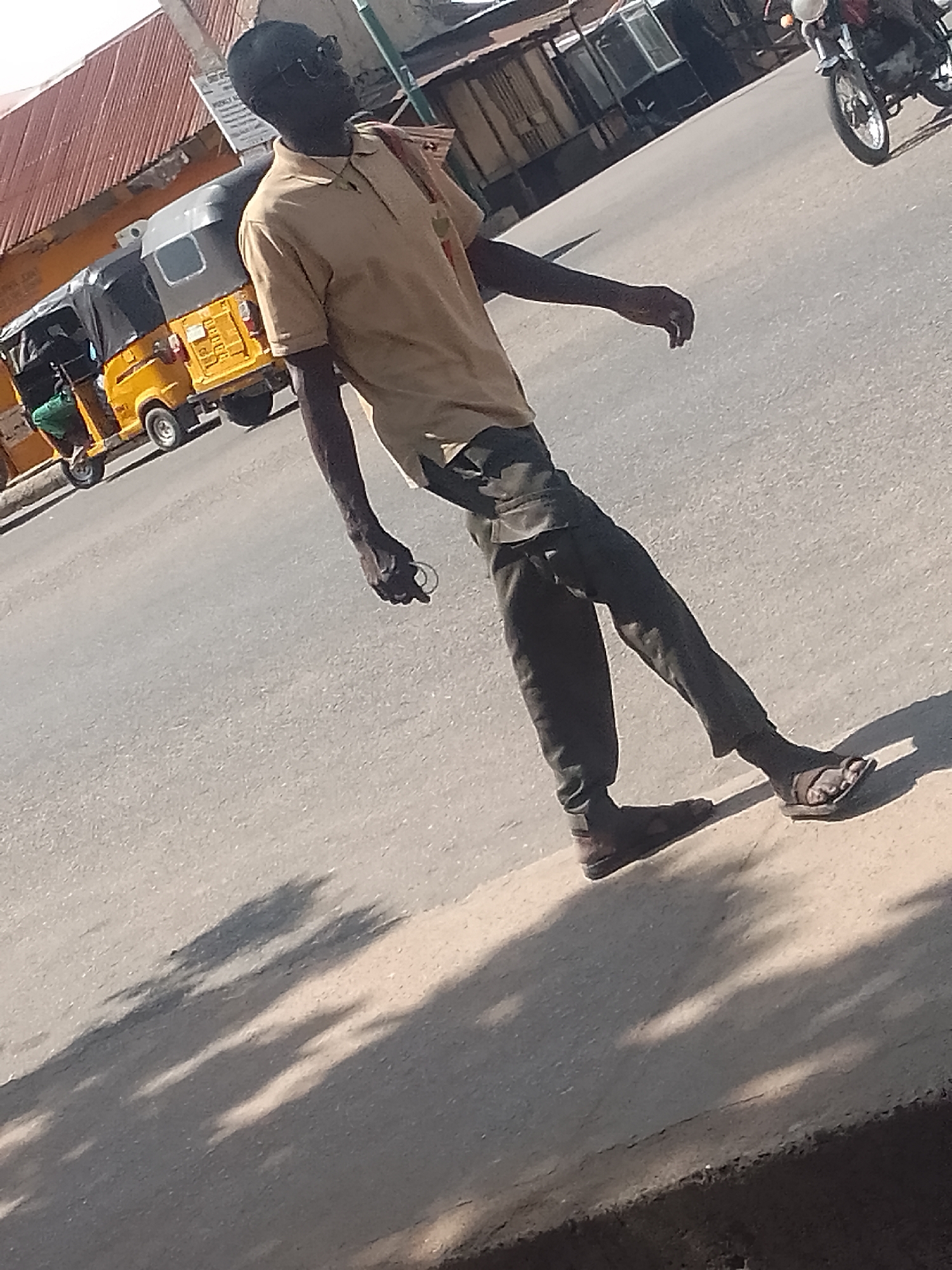 A man is depicted walking on a sunlit street, likely in an urban or semi-urban setting. He is dark-skinned, wearing dark sunglasses, a light brown short-sleeved polo shirt, and dark trousers. He has a small, light-colored, ring-like object in his left hand. His footwear appears mismatched, with a dark sandal or slip-on on his right foot and a darker, more enclosed shoe or sandal on his left.
In the background, to the man's left, a yellow tricycle taxi (often called a Keke Napep) with a black roof cover is visible. Further right in the background, a motorcycle's front wheel and part of its engine can be seen. Behind these vehicles, buildings and structures are present, including one with a reddish-brown corrugated metal roof.
The lighting indicates a bright, sunny day, with long, distinct shadows stretching across the road, suggesting either early morning or late afternoon. The presence of the Keke Napep and the general environment are consistent with the provided location context of Pantame, Nigeria. No discernible text is visible in the image.