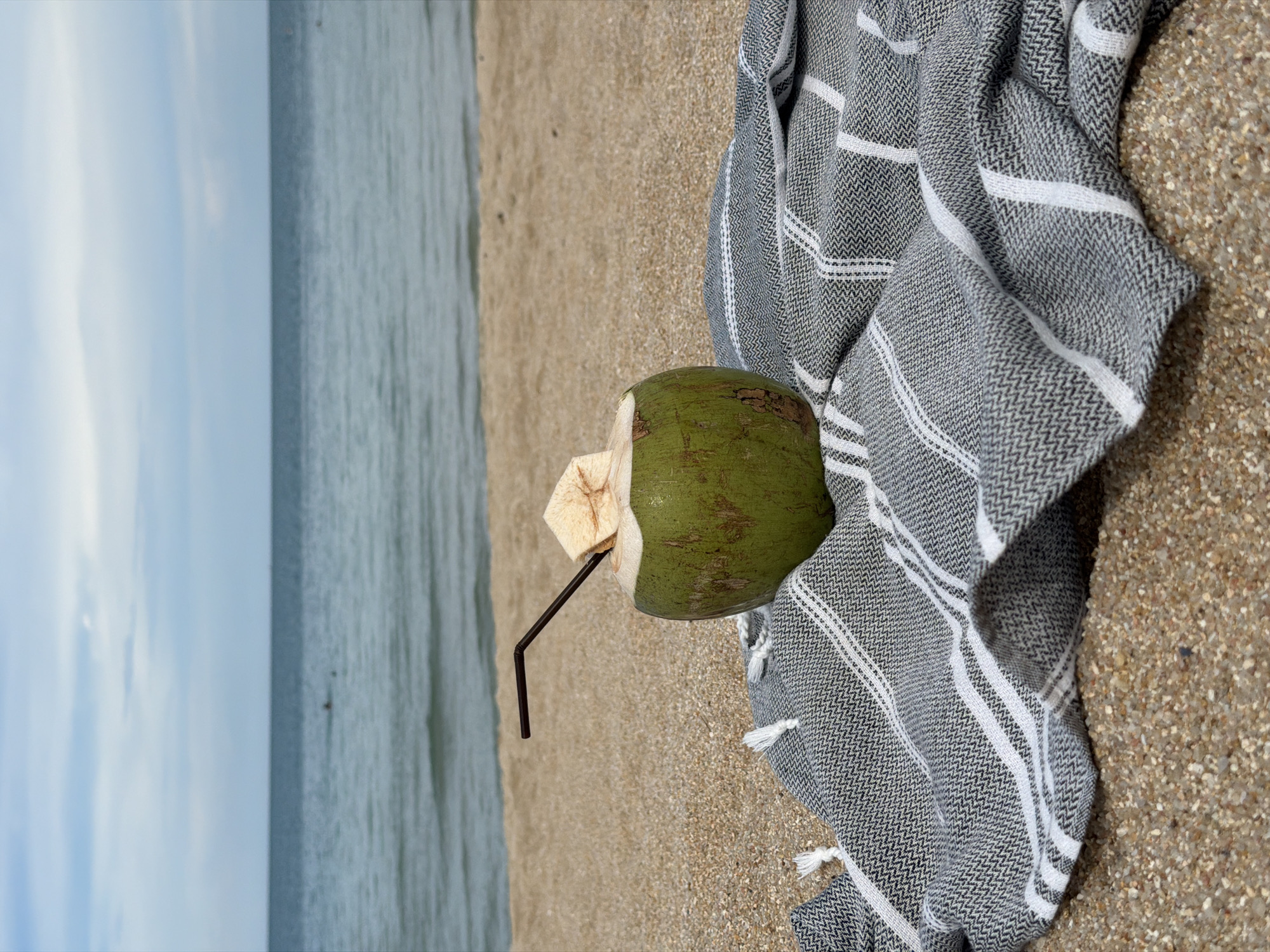 A whole green coconut with a straw and a piece of its shell acting as a cap, rests on a striped grey and white beach towel. The towel is placed on a sandy beach. In the background, the ocean meets the sky under a light blue cloudy sky. Two small figures are visible swimming in the distance. The overall scene evokes a relaxed, tropical beach setting, likely in Baan Chaweng, Thailand.