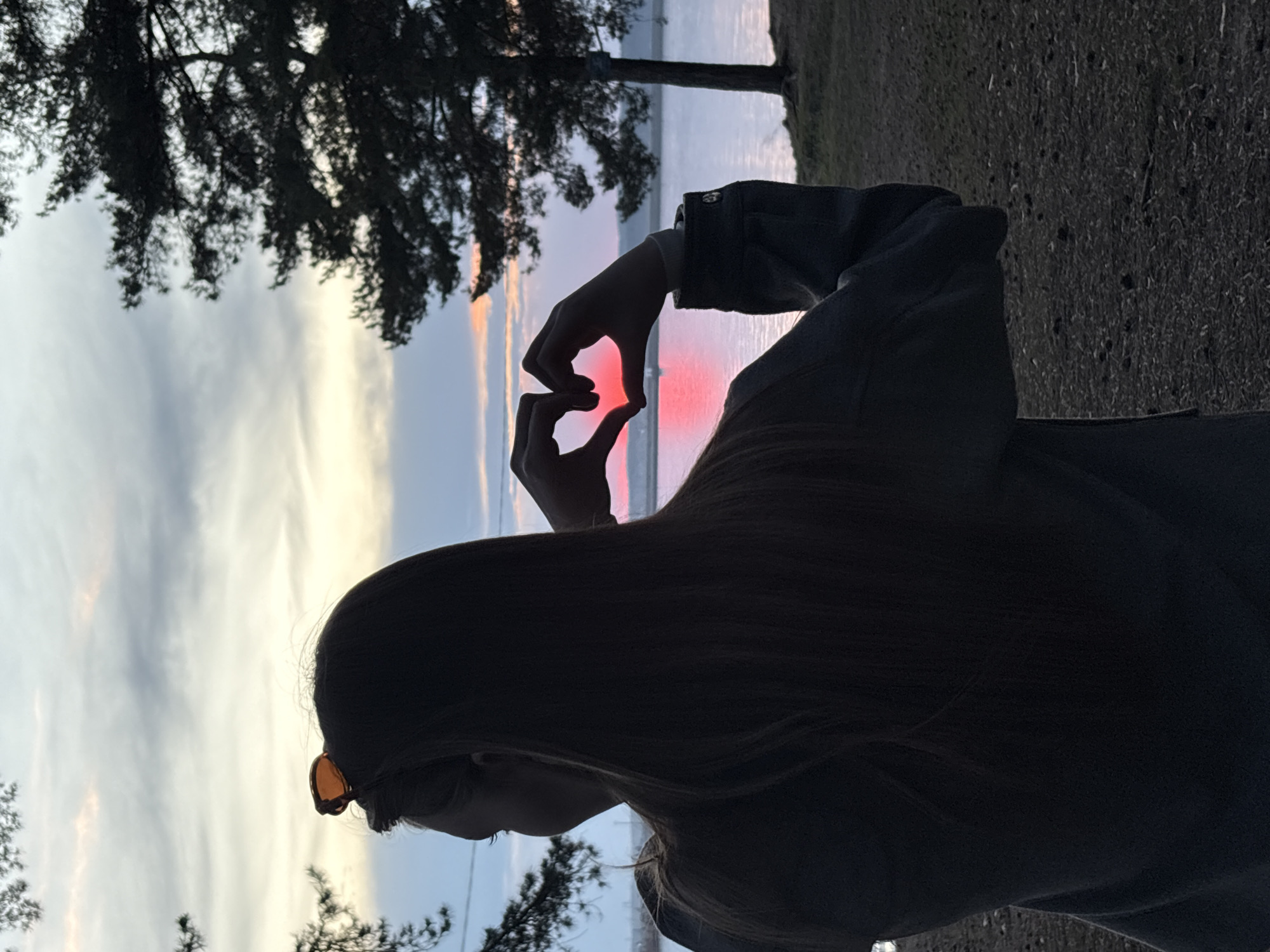 A person with long hair, wearing sunglasses, is silhouetted against a sunset or sunrise sky with water in the background. The person is holding their hands in the shape of a heart. The scene is framed by trees on either side. The location is Chaikovsky, Russia.