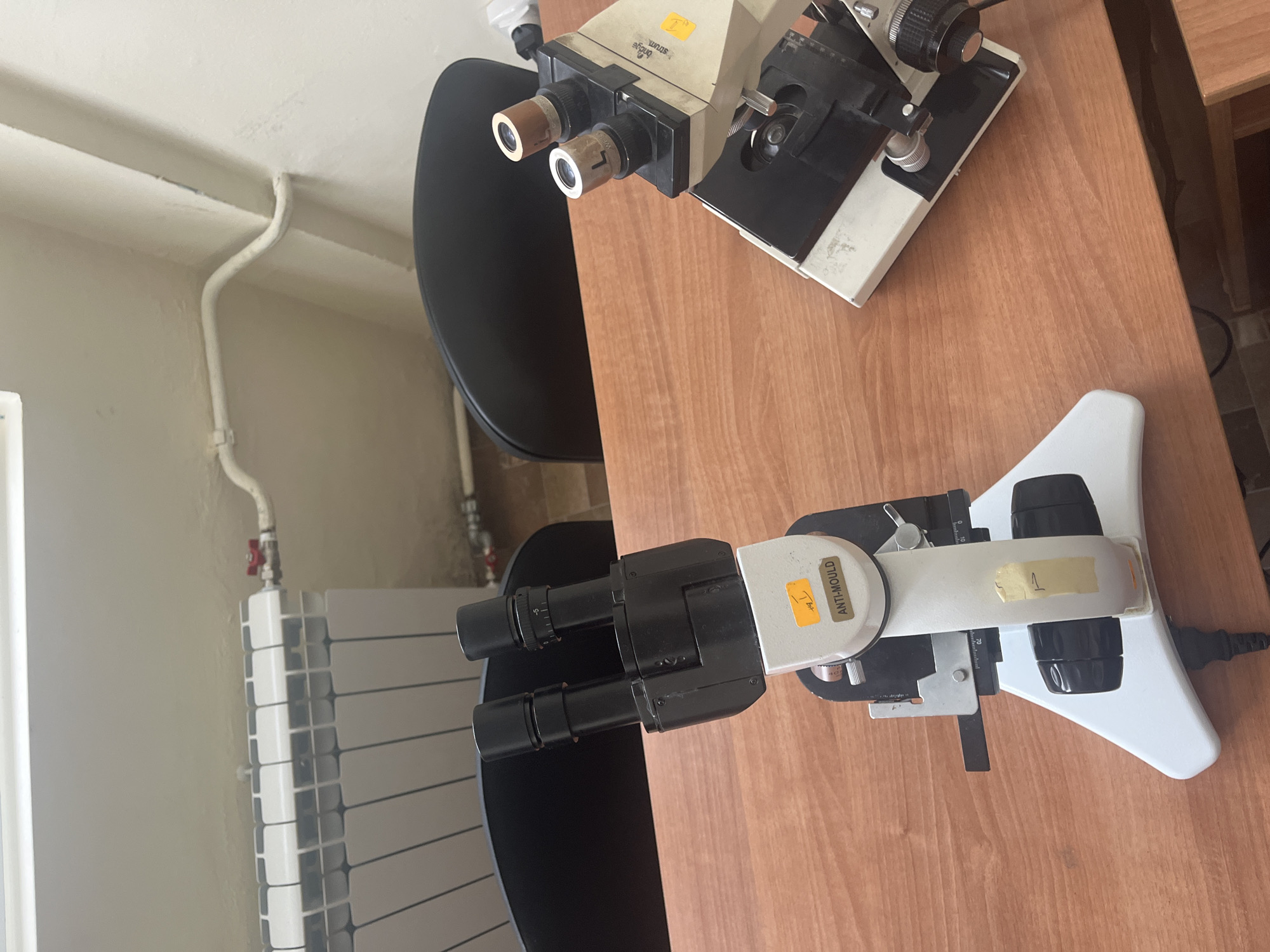 The image shows two microscopes placed on a wooden desk. One microscope is beige and black, while the other is white and black. A black chair is positioned behind the microscopes. In the background, there is a white radiator and a white wall with pipes. The location is Plovdiv, Bulgaria.