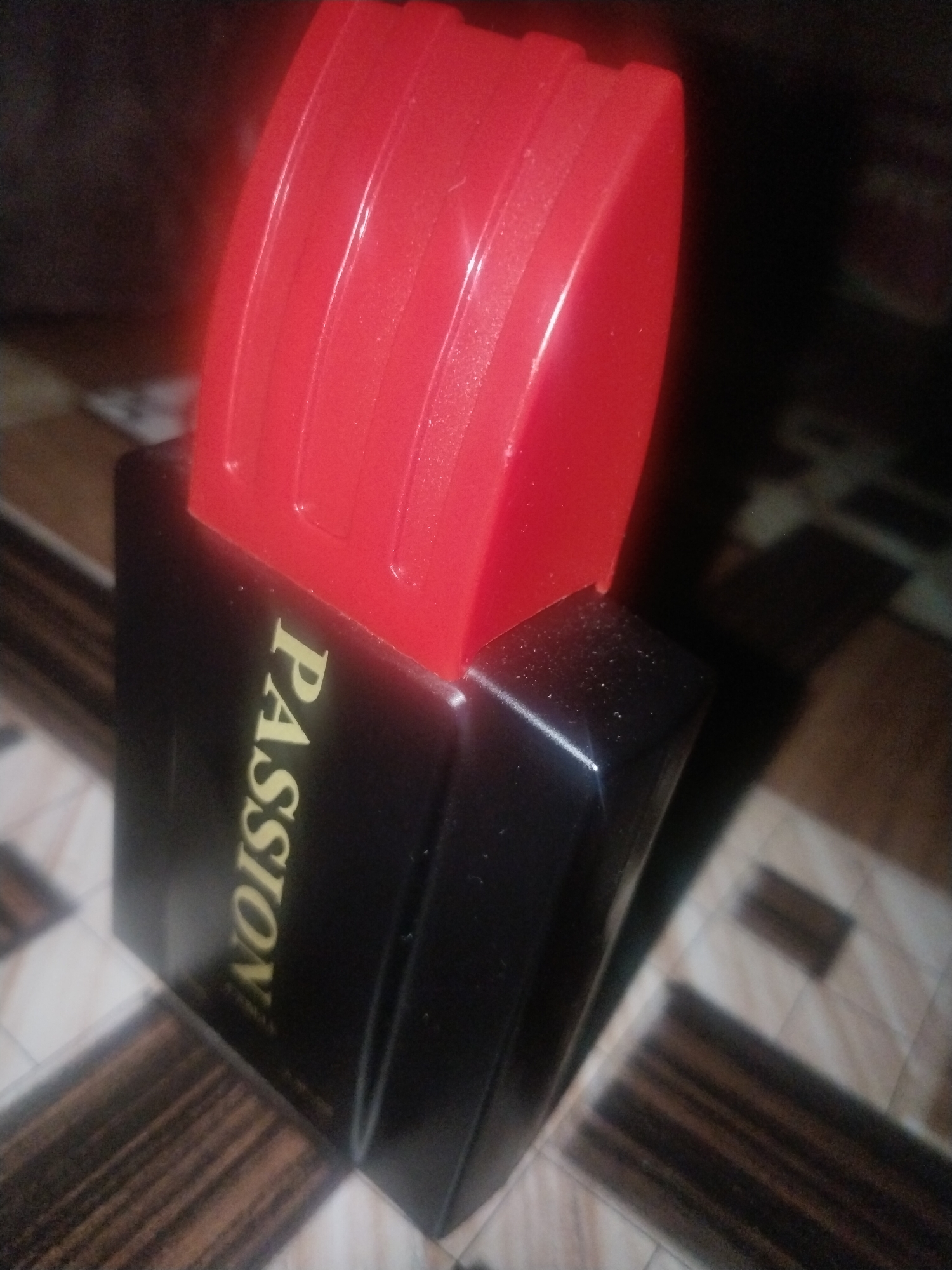 A still, medium shot depicts a bottle of "Passion" perfume. The bottle is black with the word "Passion" printed vertically in gold lettering. It has a red, curved cap. The bottle sits on a surface with a brown and white geometric pattern, likely a tiled floor. The background is dark and out of focus. There is no information available from the image to confirm the location is Uyo, Nigeria.