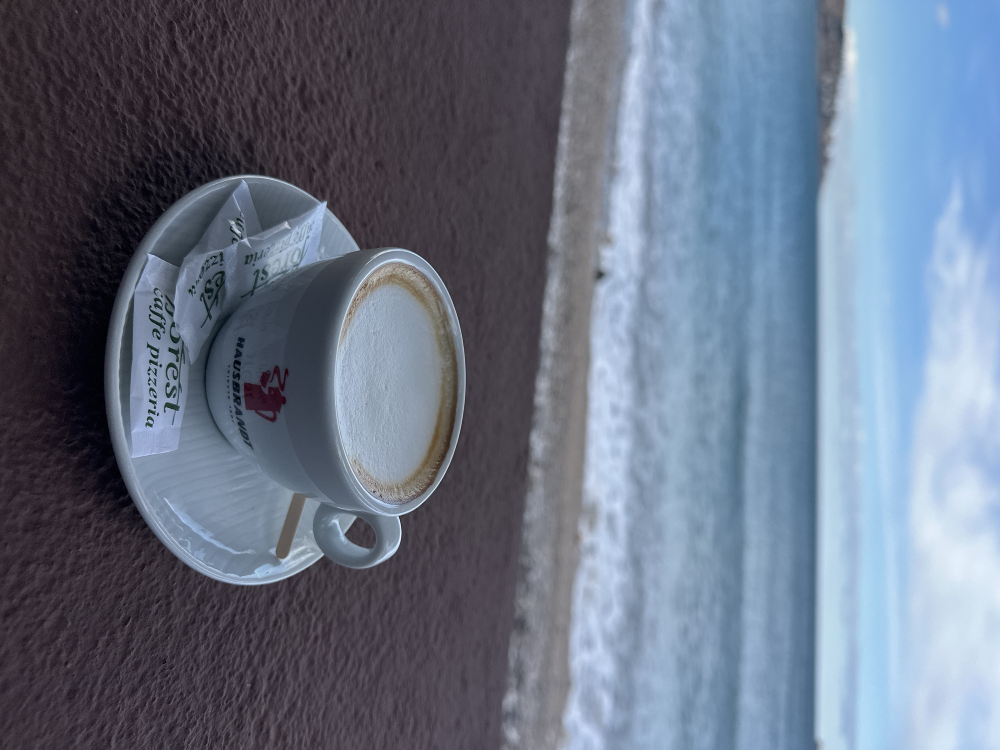 A cup of cappuccino rests on a small, white saucer with two sugar packets next to it, placed on a brown, textured surface. The cup is white with a red logo. The background shows a blurry view of the Adriatic Sea, and a beach under a partly cloudy sky. The location is plausibly a café in Bar, Montenegro, given the coastal view.