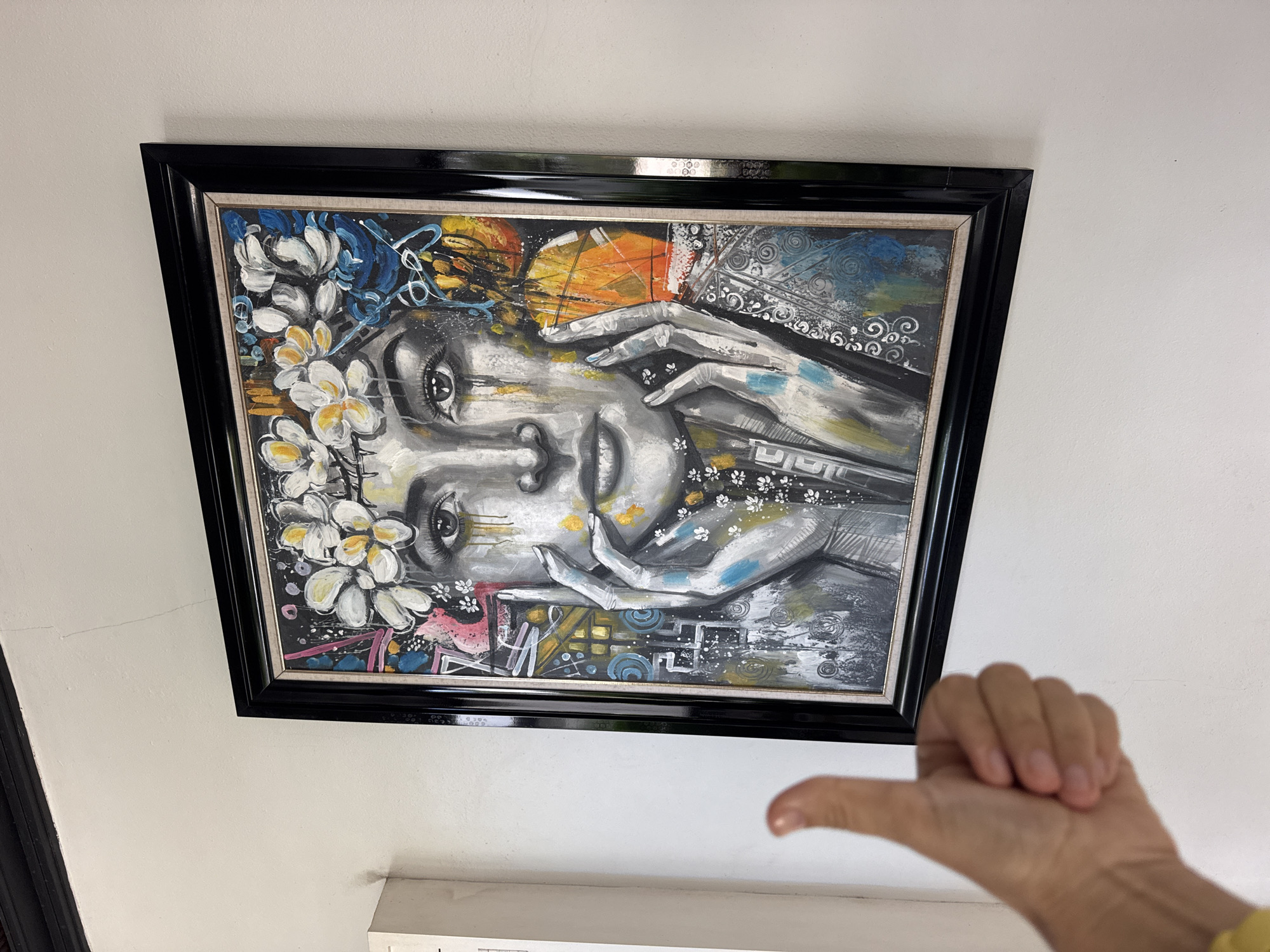 A framed painting depicting a stylized portrait of a person with hands near the face is hanging on a white wall. The painting features white flowers on the left side, and the color palette is dominated by grayscale tones with accents of blue, yellow, and orange. A hand is visible in the lower right corner of the image. The location is Kuta Selatan, Indonesia.
