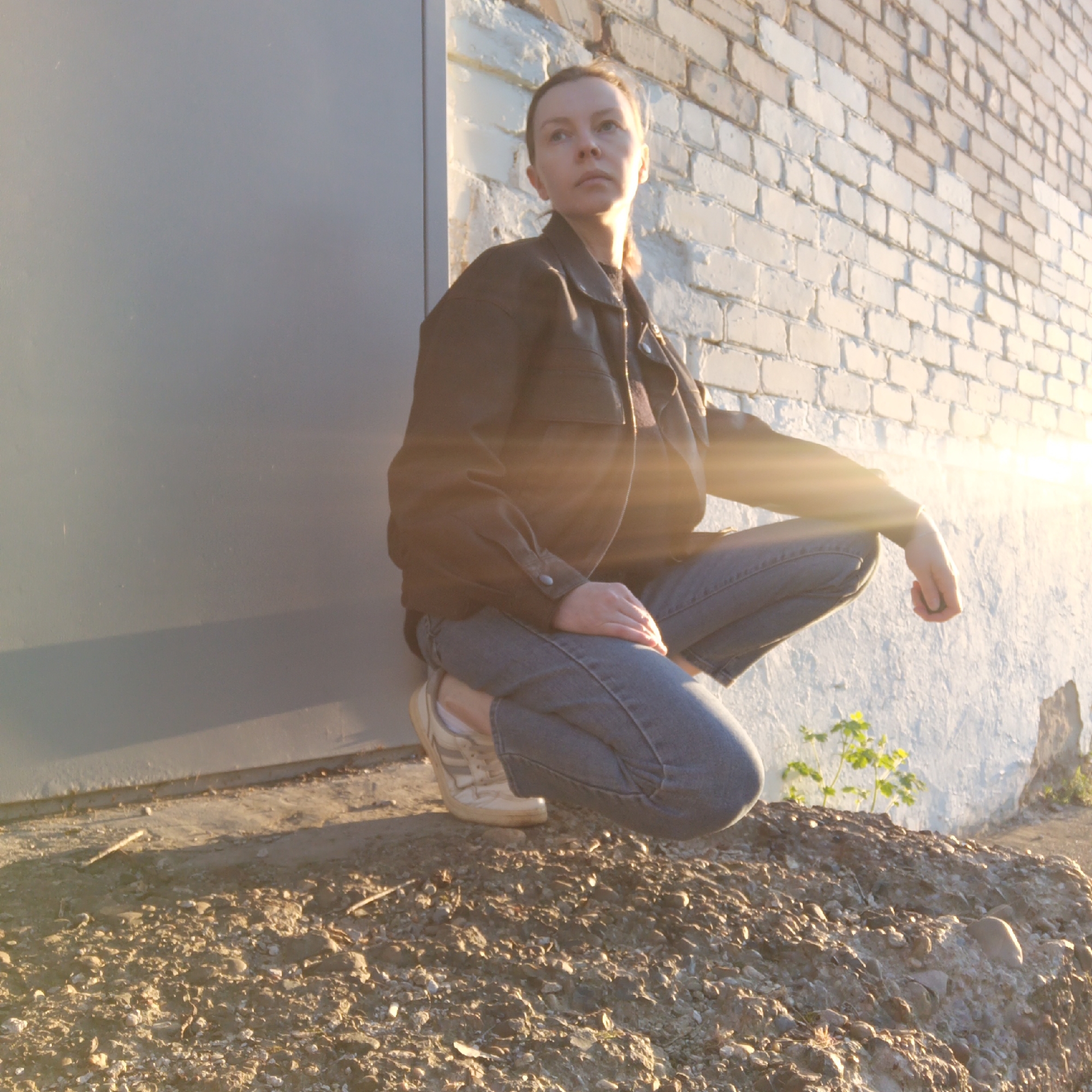 A young woman is crouched on a small mound of dirt and gravel in front of a brick wall and a gray metal door. She is wearing jeans, a black jacket, and sneakers. The sun is shining brightly, and there is some foliage growing near the wall. This photo was taken in Chaikovsky, Russia.