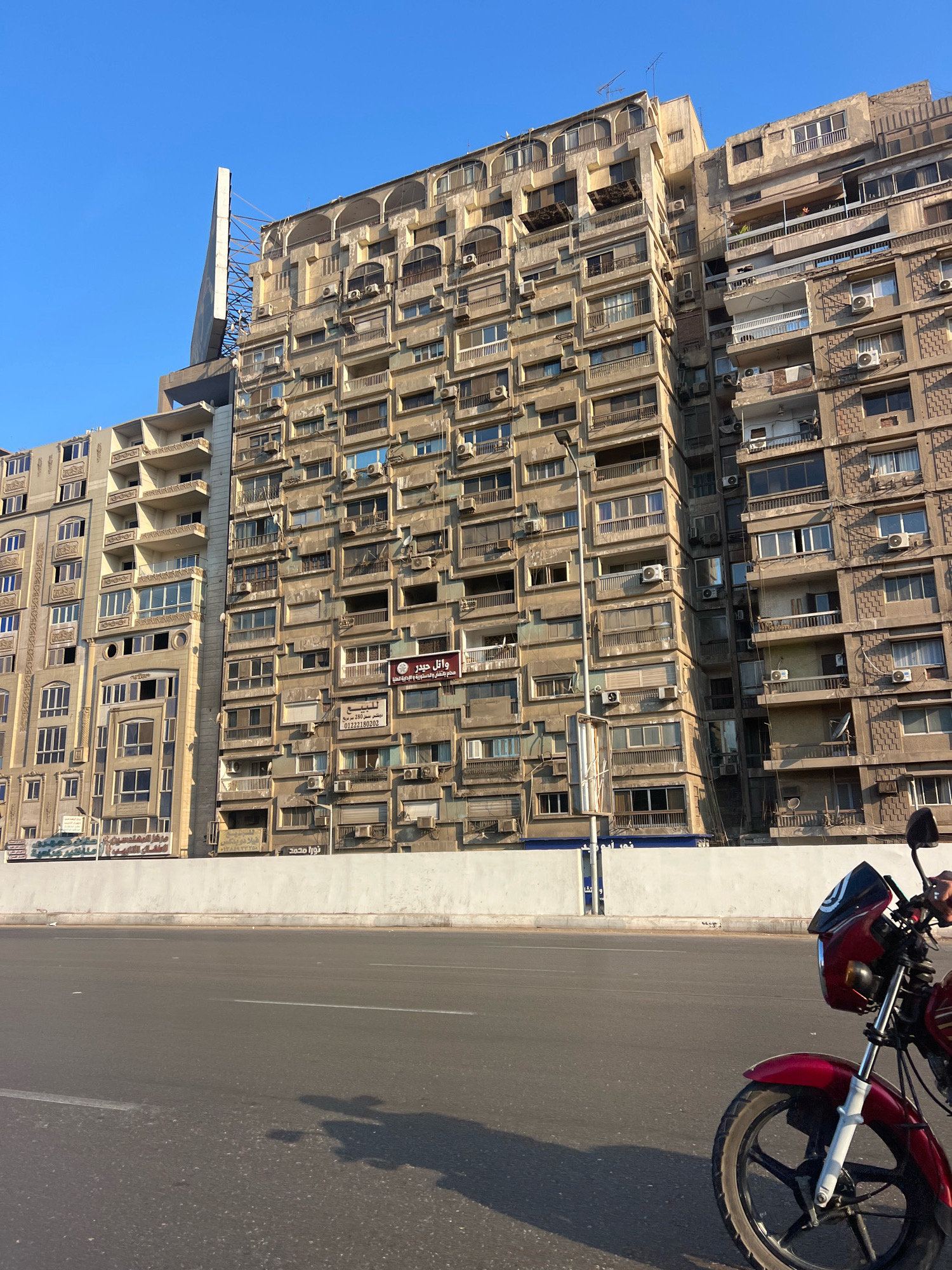 In Giza, Egypt, a street-level view captures a sunlit urban scene dominated by a massive, multi-story apartment building with a beige, weathered facade. The building exhibits a dense arrangement of windows, balconies, and air conditioning units, creating a textured and somewhat chaotic architectural style. Adjacent to this primary building is another, lighter-colored building with more regular fenestration. A large, metallic structure, possibly a billboard frame, is visible extending upwards from the top of the beige building. In the foreground, a section of a red motorcycle with a black windshield and handlebars is parked on the right side of the frame, its front wheel and part of the fender visible. The street is paved and appears to be a road with lane markings, and a white concrete barrier runs along the edge of the sidewalk, separating it from the street. The sky is a clear, bright blue.