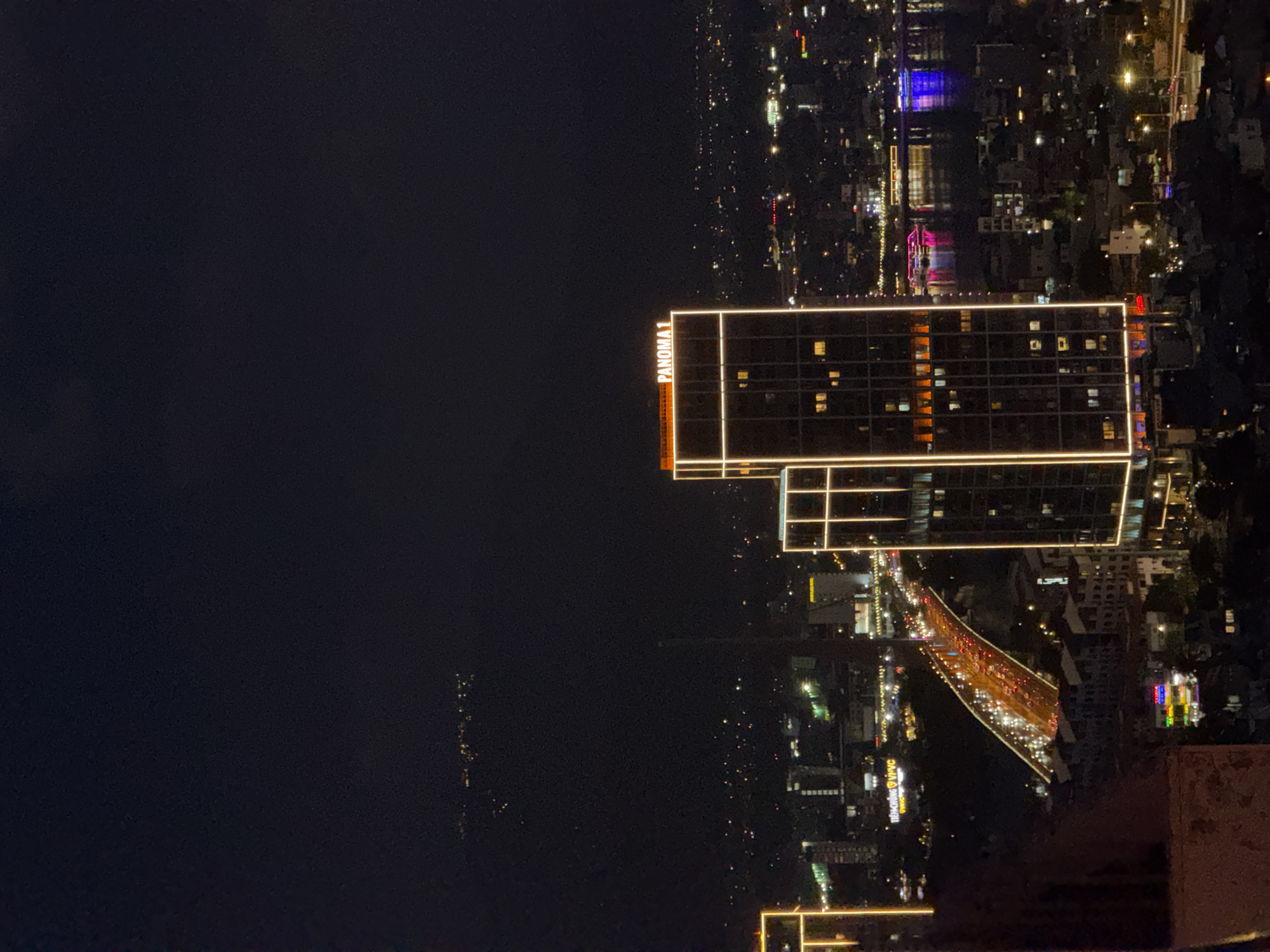 This is a nighttime aerial view of the city of Da Nang, Vietnam, focusing on the Han River and the surrounding urban landscape. The image is dominated by the dark night sky, with scattered city lights below creating a glittering effect.
Prominently featured are two modern skyscrapers with illuminated outlines, one of which displays the text "PANOMA 1" in orange neon letters across its top. These buildings are indicative of the city's modern architectural development.
A long, elevated bridge, likely part of the city's infrastructure, spans across the frame. It is brightly lit with a continuous stream of car headlights and taillights, suggesting a busy traffic flow. The bridge's lights reflect on the dark water of the Han River below.
Along the riverbanks, a variety of buildings and lights are visible, ranging from commercial establishments to residential areas. Several boats or vessels are also seen on the river, some with colorful lights, adding to the vibrant nighttime atmosphere. In the distance, a faint line of lights suggests further urban sprawl or perhaps a more distant part of the city or coastline.
The foreground shows a dark, textured surface, possibly a building facade or a rooftop edge, from which the photo is taken, providing a sense of perspective and height. The overall scene captures the dynamic and illuminated character of Da Nang at night.