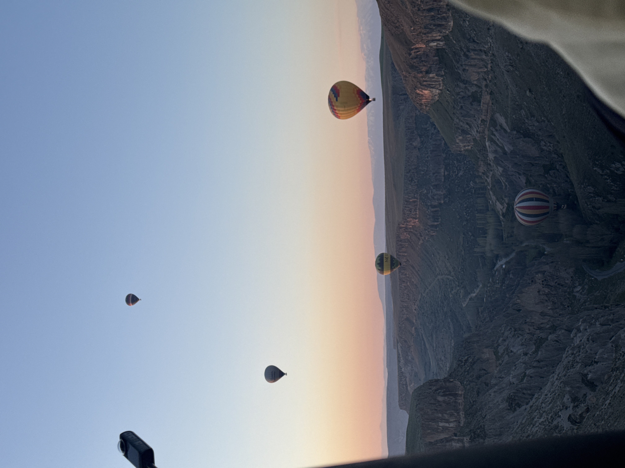 The image presents a breathtaking aerial view of multiple hot air balloons drifting over a majestic canyon landscape, likely during sunrise or sunset due to the soft, gradient light. The sky, filling the upper and left sections of the frame, transitions smoothly from a clear, pale blue at the top to warm hues of peach, orange, and pink towards the horizon on the right.
Below and to the right, a vast and rugged canyon system dominates the scene. It features steep, dark rock formations, intricate cliff faces, and scattered vegetation, with a river or body of water snaking through the canyon floor. Distant hills or mountains line the horizon.
At least five hot air balloons are visible. A prominent yellow balloon with red, blue, and purple stripes floats against the colorful horizon on the right. Below it, closer to the canyon, another balloon with red and white stripes is partially obscured by shadows. Higher up and further left, a smaller, dark-colored balloon is silhouetted against the blue sky. Another black balloon with indistinct white text is seen in the mid-left sky, while a dark green and yellow balloon, with faint text that appears to include "NES", is positioned near the canyon rim on the right.
The photograph is taken from within another hot air balloon, with parts of its structure visible as dark edges at the top and bottom of the frame. A black camera or recording device is mounted in the foreground at the bottom left. The weather appears clear and calm, ideal for ballooning. While no individuals are visible, the presence of multiple balloons signifies an organized event or tour. The scene evokes a sense of grandeur and peaceful adventure, set against the dramatic backdrop of Yeşilhisar, Turkey.
