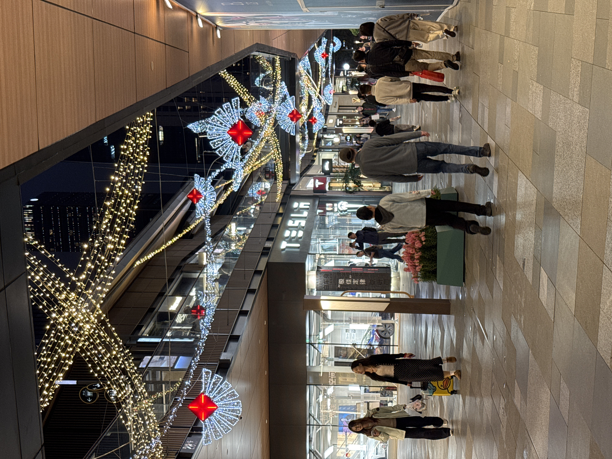 The image shows a pedestrian area in what appears to be an outdoor shopping mall or commercial district. The location is likely Jinjiang District, China, based on the presence of Chinese characters on signage.
Several people are walking along the pedestrian walkway. Many of them are wearing casual attire. There are groups of people in different parts of the walkway.
The area is decorated with Christmas-style lighting, including strings of lights and illuminated starburst decorations with red centers.
Buildings are visible on the left side of the image. One building has "TESLA" signage. The pavement is made of large, rectangular tiles. Planter boxes with flowers are also visible.