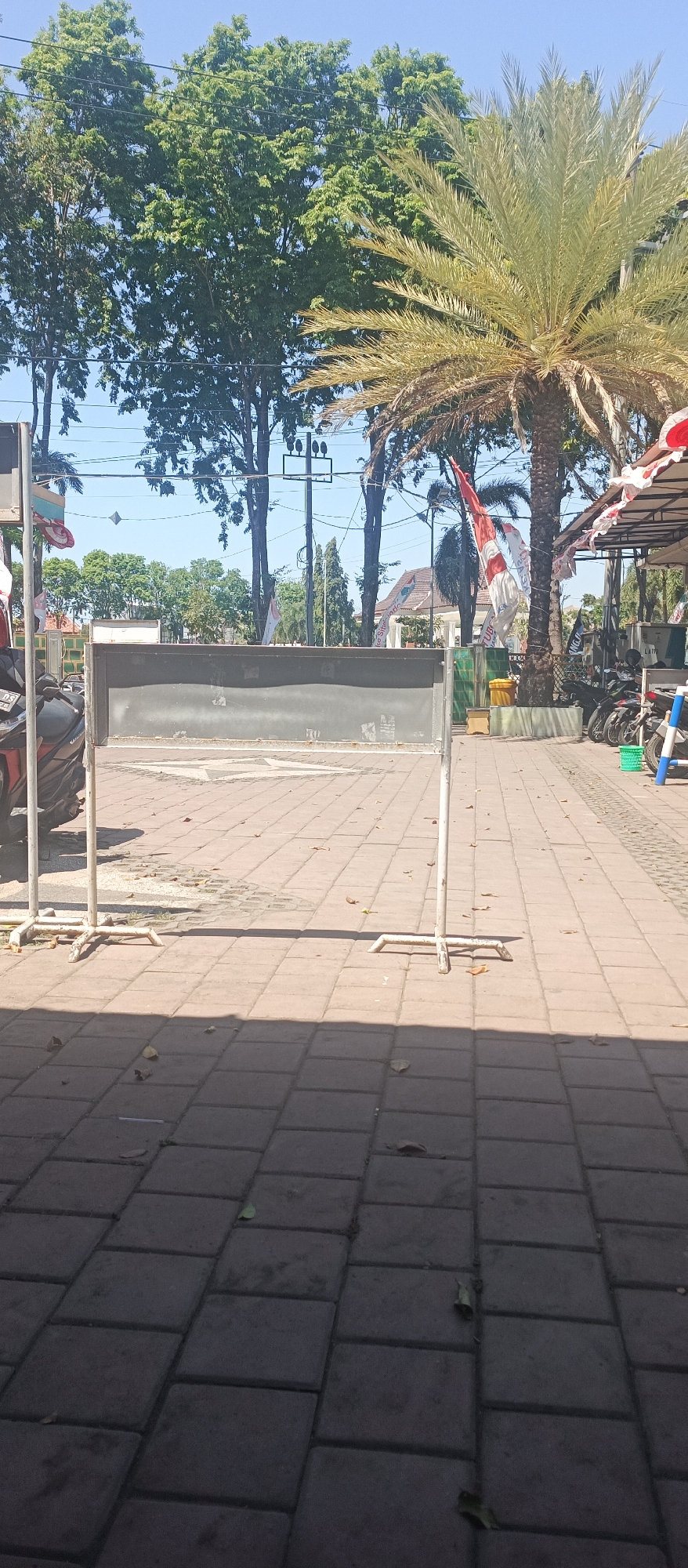 The image depicts an outdoor scene on a sunny day, likely in Situbondo, Indonesia. A large, dark rectangular sign, possibly a temporary barrier or information board, is centrally positioned in the frame. It is supported by two white metal stands with cross-shaped bases. The ground is paved with reddish-brown rectangular bricks, and scattered brown leaves are visible on the paving stones.
In the background, a tall palm tree with prominent fronds dominates the right side of the image. Behind the sign and palm tree, several leafy green trees stand against a clear blue sky. Some Indonesian flags, red and white, are visible, one prominently hanging from a pole near the palm tree and another with white tassels dangling from an awning. Power lines and poles are also present in the background.
To the left of the sign, a portion of a red and black motorcycle is visible. To the right, under a canopy or awning with red and white decorative tassels, several parked motorcycles are lined up. A yellow trash bin is also located near the parked motorcycles. The overall impression is of a public outdoor area, possibly a plaza or walkway, decorated for an event.