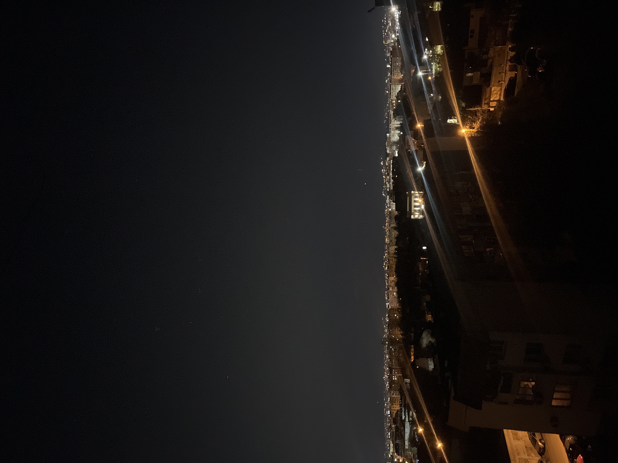 An aerial view at night in Sofia, Bulgaria. The right portion of the image depicts lit buildings and a road or street, with light beams emanating from street lights. The left portion of the image consists of a dark, night sky. No people are visible.