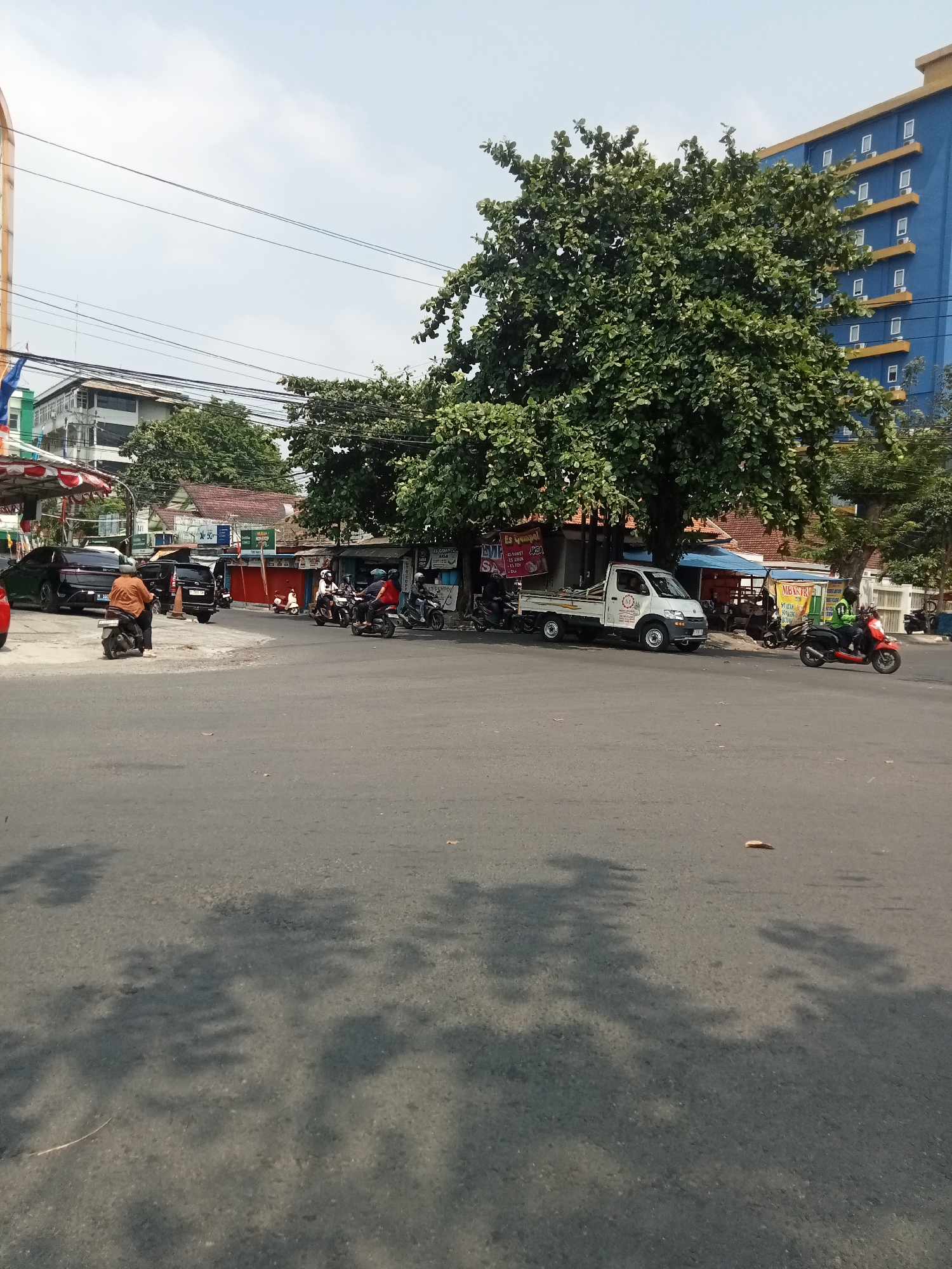 The image captures a street scene in Semarang, Indonesia, likely during daylight. The foreground is dominated by an asphalt road with distinct tree shadows cast across it, suggesting the presence of large trees nearby. In the mid-ground, a busy intersection is visible. Several motorcycles are present, with some riders wearing helmets, indicating ongoing traffic. A white pickup truck is parked on the side of the road, possibly at a small market or roadside stall area. Various small buildings with awnings and signage line the street, some appearing to be shops. A large, leafy green tree stands prominently in the center of the frame, providing shade. In the background, a modern blue and yellow multi-story building rises above the smaller structures, contrasting with the older architecture. The sky is overcast with visible power lines crisscrossing the frame, a common feature in urban Indonesian landscapes. A few individuals are visible, including a person on a motorcycle wearing an orange shirt and a helmet, and other figures on motorcycles in the background. The overall impression is one of a lively, functional urban environment.