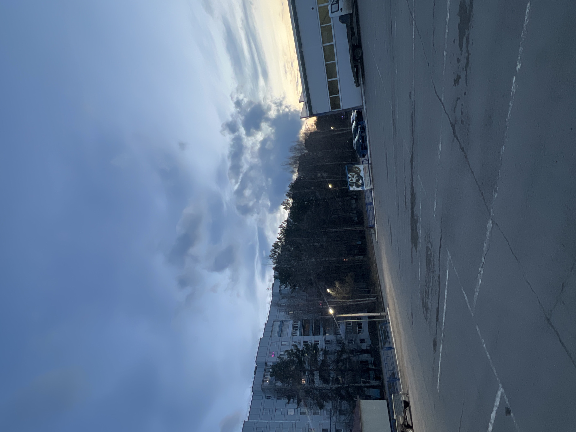 The image depicts an urban or suburban scene in Chaikovsky, Russia, likely at dusk or dawn. The overall impression is quiet and somewhat cool-toned, with artificial lights beginning to contend with the natural light.
The sky dominates the upper portion of the frame, filled with a dramatic interplay of dark, heavy clouds and lighter, elongated clouds. A warm, golden glow on the horizon, possibly from a setting or rising sun, peeks through the cloud cover.
In the midground, a prominent, multi-story grey apartment building stands on the left, with numerous windows, some of which are illuminated, suggesting occupancy. A dense line of tall, dark trees runs horizontally across the middle of the scene, obscuring parts of the buildings and adding a natural element. Several streetlights are visible among the trees and along the asphalt area, with at least one clearly illuminated, casting a yellow light.
The foreground features a wide asphalt surface, which appears to be a parking lot or roadway. It has distinct white painted lines marking parking spaces or lanes. Scattered dark, wet patches on the asphalt suggest recent rainfall.
On the right side of the image, beyond the trees and parking area, there is a long, white commercial or industrial building with a series of rectangular windows. A white truck is parked near this building.
Near the center-right, a white rectangular sign is visible. It features some indistinct graphics, possibly related to food, and some text. The most discernible part of the text appears to be "3C" or "ЭС" (Russian letters), with other smaller text below it remaining illegible.
No people are visible in the scene. The presence of both residential and commercial/industrial structures, along with the parking lot and trees, suggests a mixed-use area of the city.