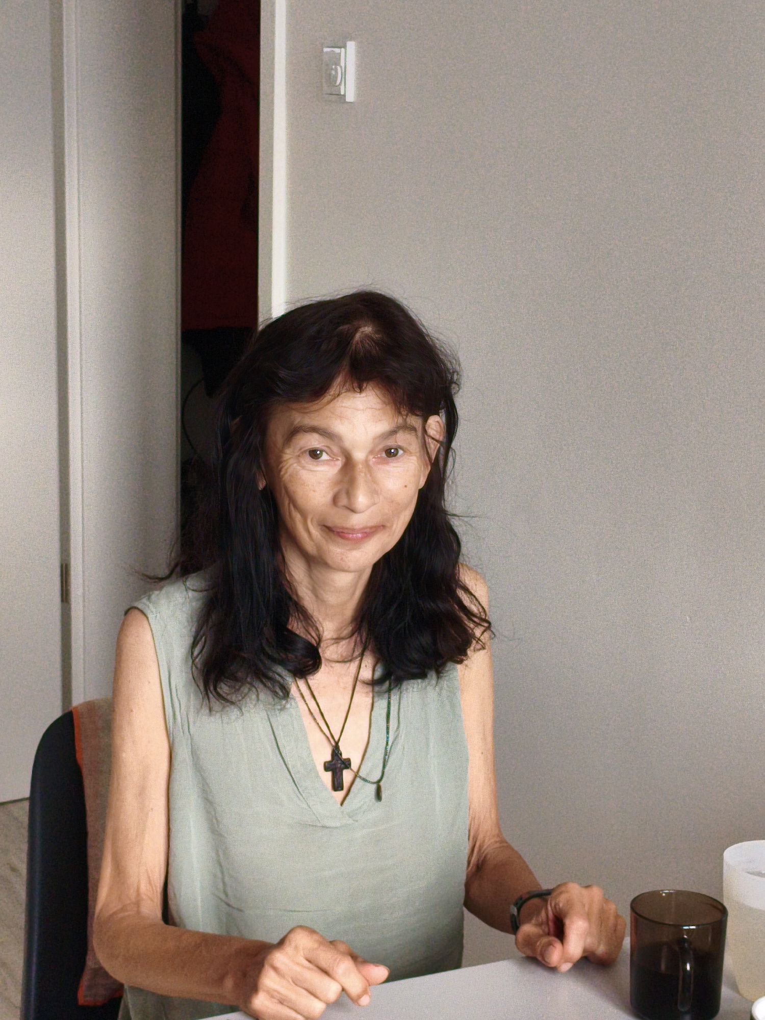 A woman with dark, wavy hair is seated at a table, facing the camera. She is wearing a light green sleeveless top with a V-neck and a dark cross pendant on a necklace. Her left arm rests on the table, and her right hand is slightly extended. To her right, a dark glass mug and a white cylindrical object are visible on the table. The background consists of a plain gray wall and a partially visible doorway with a red garment hanging inside. The scene appears to be indoors, possibly in a home or a quiet establishment. The woman is looking directly at the viewer with a slight smile. The lighting is soft and diffused. There is no specific indication of the city of Wolgast, Germany in the image.