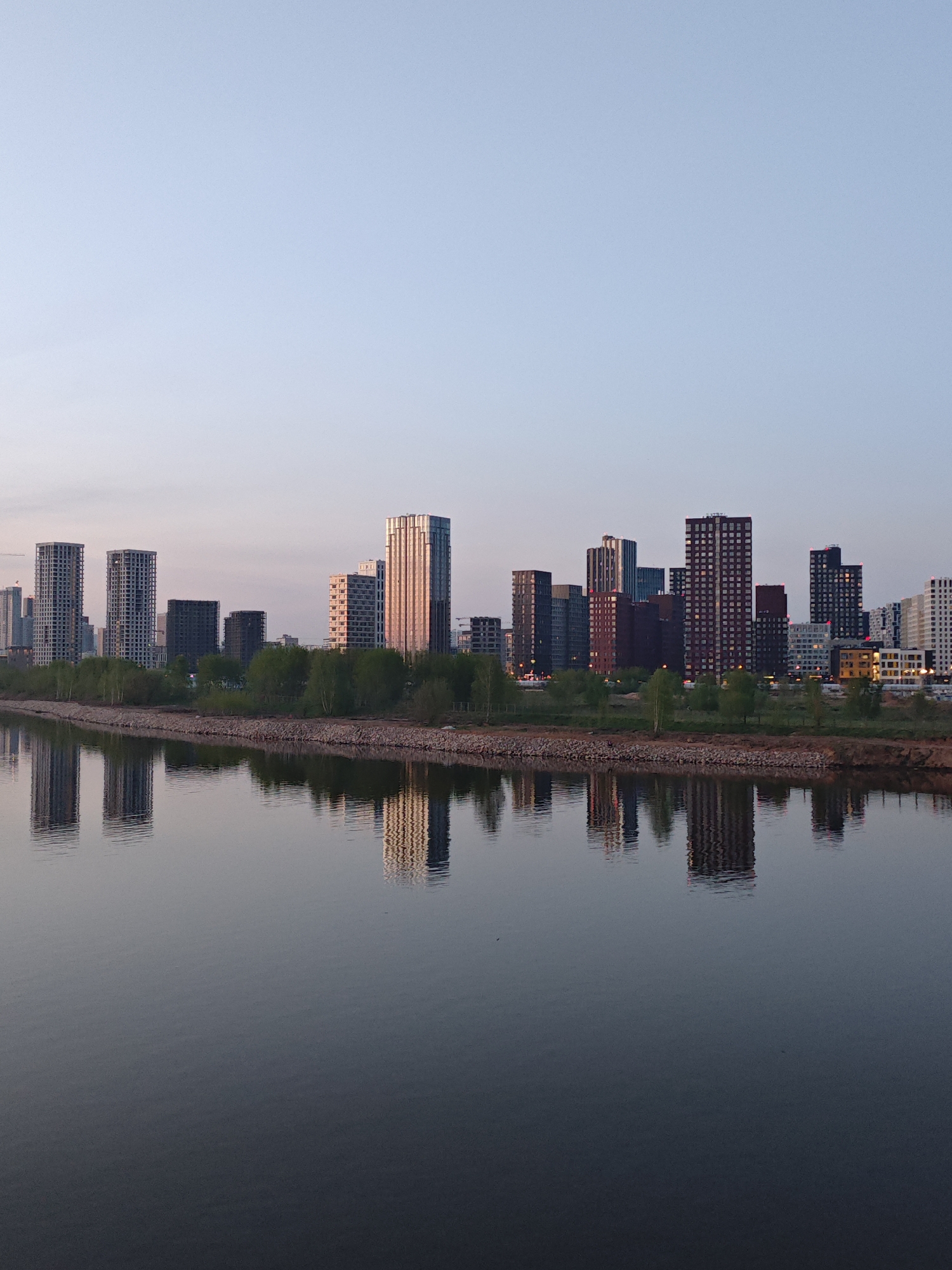 The image depicts a cityscape reflected in a body of water. In the foreground, the water surface is calm with blurred reflections of the buildings. The midground shows a shoreline with vegetation and a row of rocks or embankment. The background features a skyline of modern buildings and skyscrapers, some with reflective surfaces. The sky is a gradient of light blue and pink, suggesting either sunrise or sunset. The location is Unknown, Unknown. There are no people visible in the image.