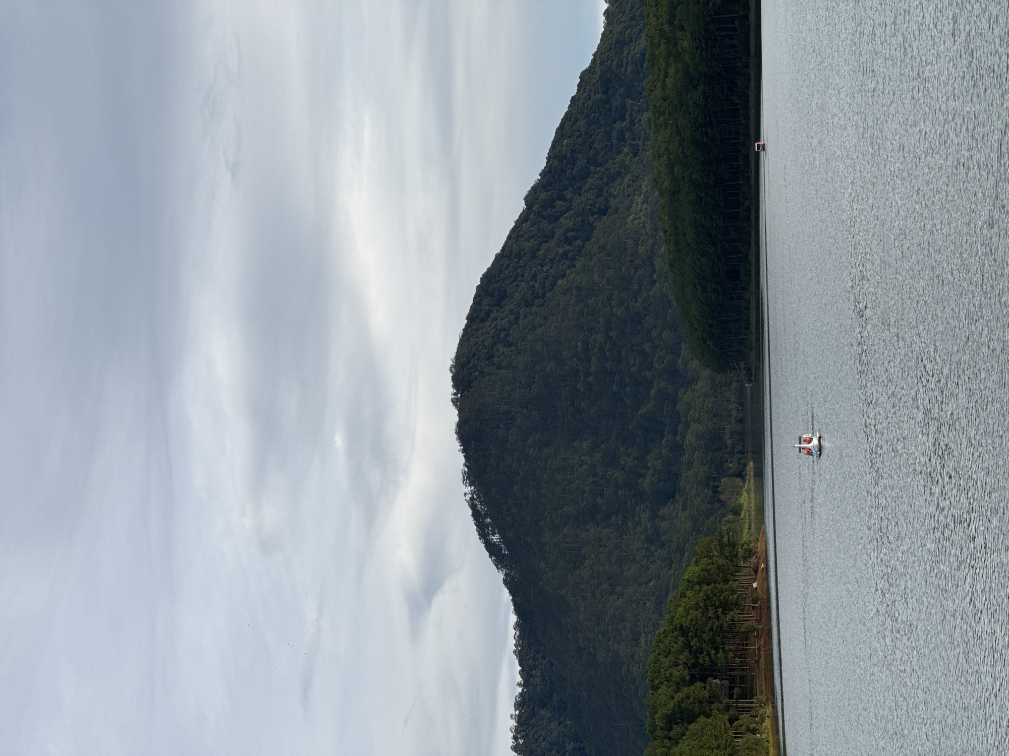 A slightly rotated scenic shot captures a lake scene at Tuyen Lam Lake, Da Lat, Vietnam. Predominantly visible is a large body of water with visible surface ripples. The lake is flanked on one side by a tree-covered mountain range, which takes up a substantial portion of the view. In the lake, there is a paddle boat with some indistinct figures. The sky is covered with many clouds and appears overcast. There is a patch of dense forest along the lake's edge.