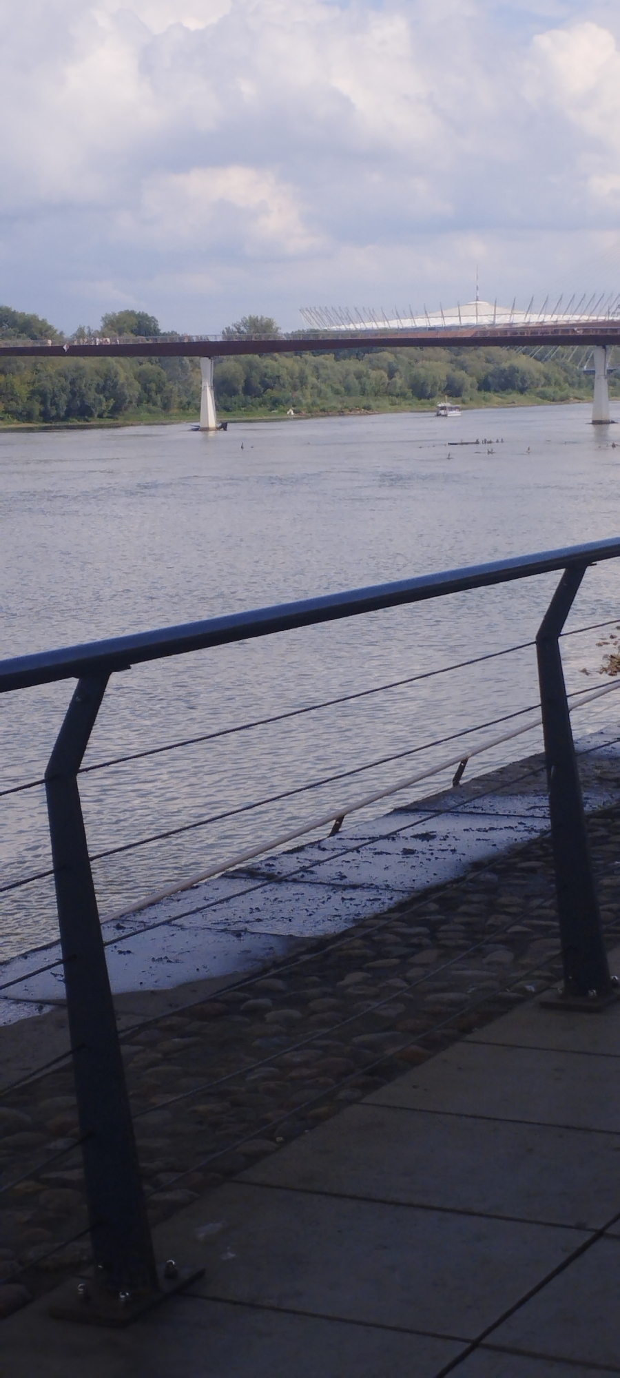The image depicts a view of the Vistula River in Warsaw, Poland, from a pedestrian walkway. In the foreground, a dark gray metal railing with thin wires runs horizontally across the frame. The walkway itself is paved with square concrete slabs and bordered by a textured area of small, rounded stones. The Vistula River is calm and reflects the cloudy sky. In the middle ground, a line of trees forms a green bank on the opposite side of the river. A modern white bridge with a distinctive curved structure is visible in the background, spanning the river and connecting to land beyond the trees. Several small boats and what appear to be individuals on paddleboards are scattered on the water. The sky is filled with white and gray clouds.