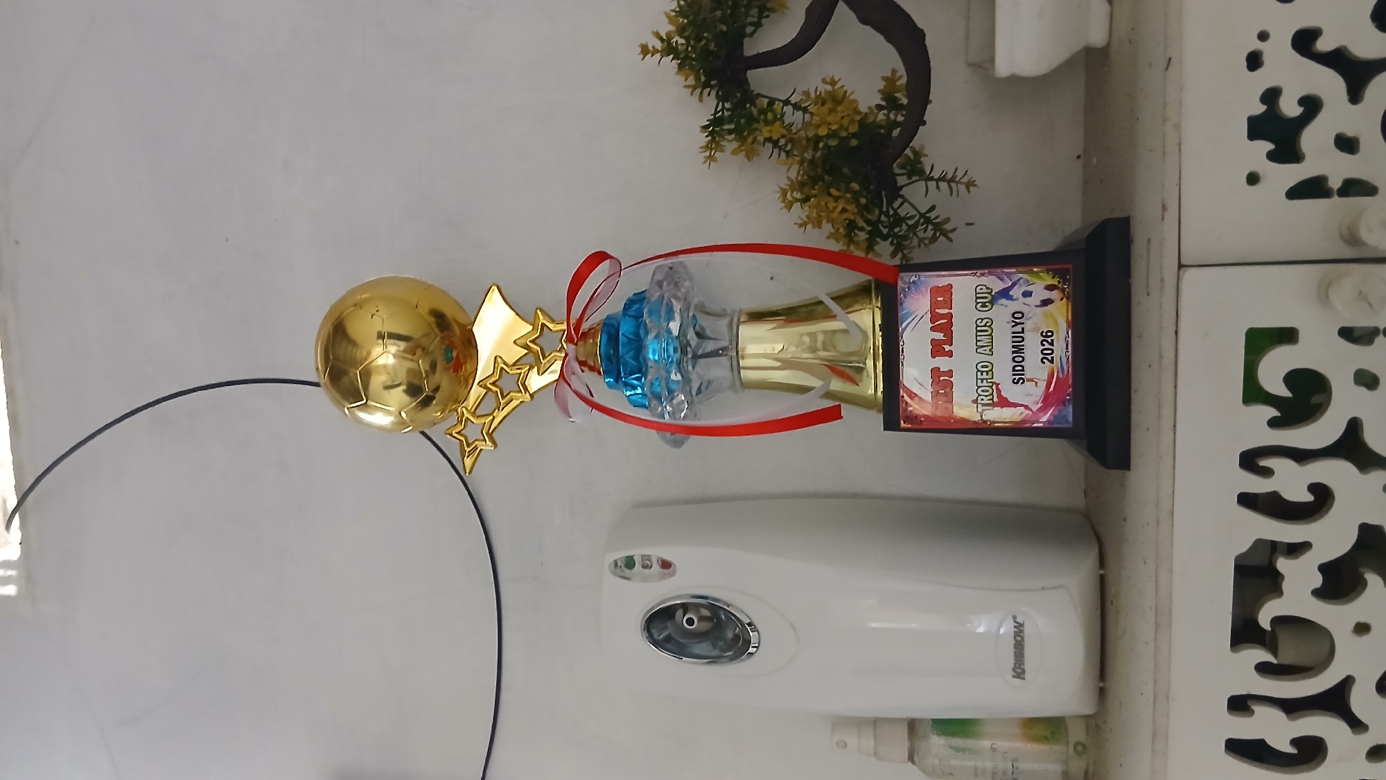 In Klambir Lima Kebun, Indonesia, a trophy for "BEST PLAYER TROfeo Amus Cup SIDOMULYO 2026" stands on a white shelf. The trophy is gold with a blue and clear crystal base, adorned with red and white ribbons. To the left of the trophy is a white automatic air freshener, labeled "Krisbow". A small spray bottle is partially visible to the left of the air freshener. To the right of the trophy, a decorative plant with yellow leaves is situated. A decorative white cabinet with an ornate cutout pattern is below the shelf. The background is a plain, light-colored wall. A thin black wire is visible extending from the top left corner of the frame, across the wall.