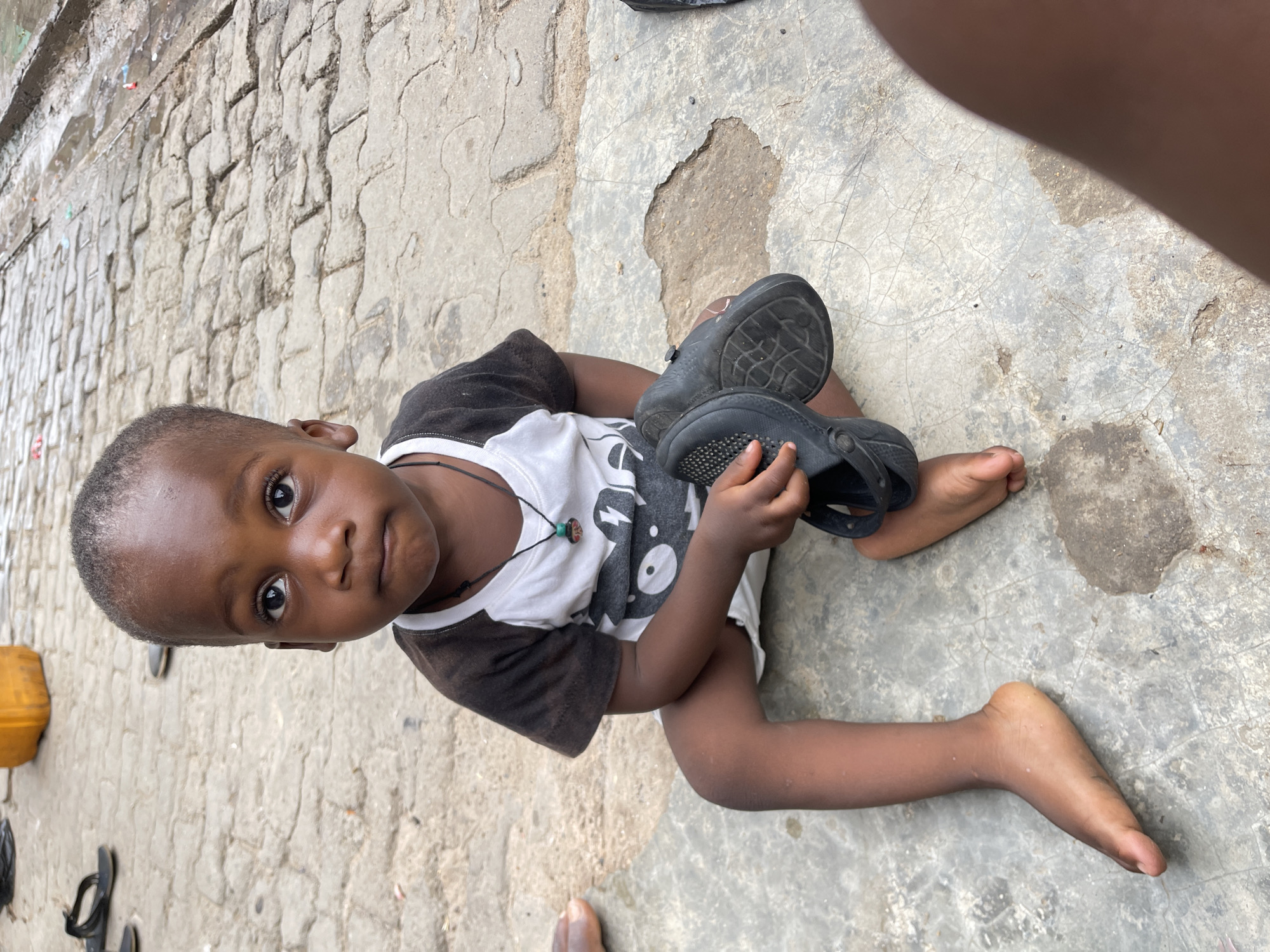 A young child, likely a boy, with dark skin and short, closely cropped hair, is seated on a worn, cracked concrete ground in an outdoor setting in Nyanya, Nigeria. He is looking directly at the camera with a calm and observant expression.
The child is wearing a white t-shirt with black short sleeves and a graphic design on the chest, which includes a lightning bolt shape. A dark beaded necklace is visible around his neck. He is holding a pair of black clogs or sandals in his hands, with their patterned soles visible.
The ground beneath him is a mix of textures: smooth concrete with fine cracks and some areas resembling rougher pavement or cobblestones, particularly towards the left of the frame. In the background, to the far left, a bright yellow jerry can is visible, alongside what appear to be other dark sandals or shoes on the ground. Partial views of another person's leg and foot are visible in the top right and bottom left corners of the frame. The scene is brightly lit, indicating it is daytime. There is no discernible text visible on any objects or clothing.