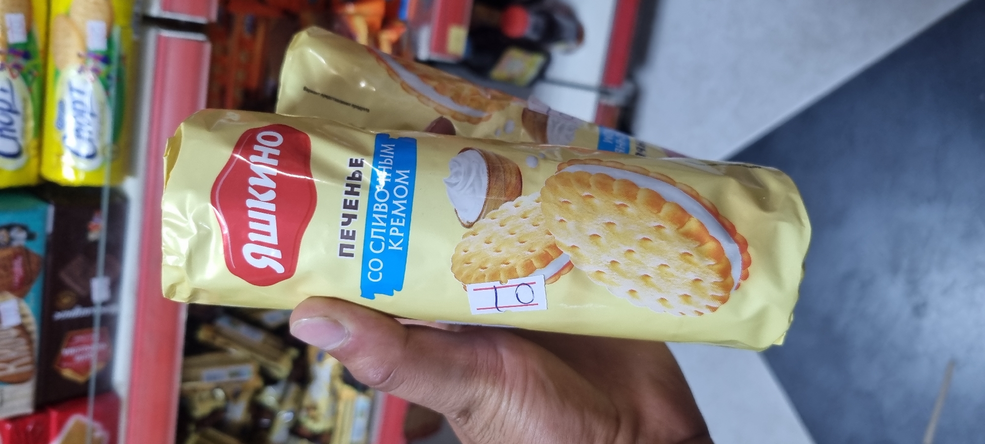 A person's hand holds a yellow package of cookies in a store aisle. The package features a red logo with Cyrillic text that appears to read "ЯШКИНО" (Yashkino), followed by smaller text in blue stating "ПЕЧЕНЬЕ СО СЛИВОЧНЫМ КРЕМОМ" (Biscuits with Cream Filling). Two cream-filled sandwich cookies are depicted on the front of the package. A small white sticker with handwritten "10" is affixed to the lower left of the packaging. In the blurred background, shelves are stocked with various food items, suggesting a retail environment in Ismoili Somoni, Tajikistan.