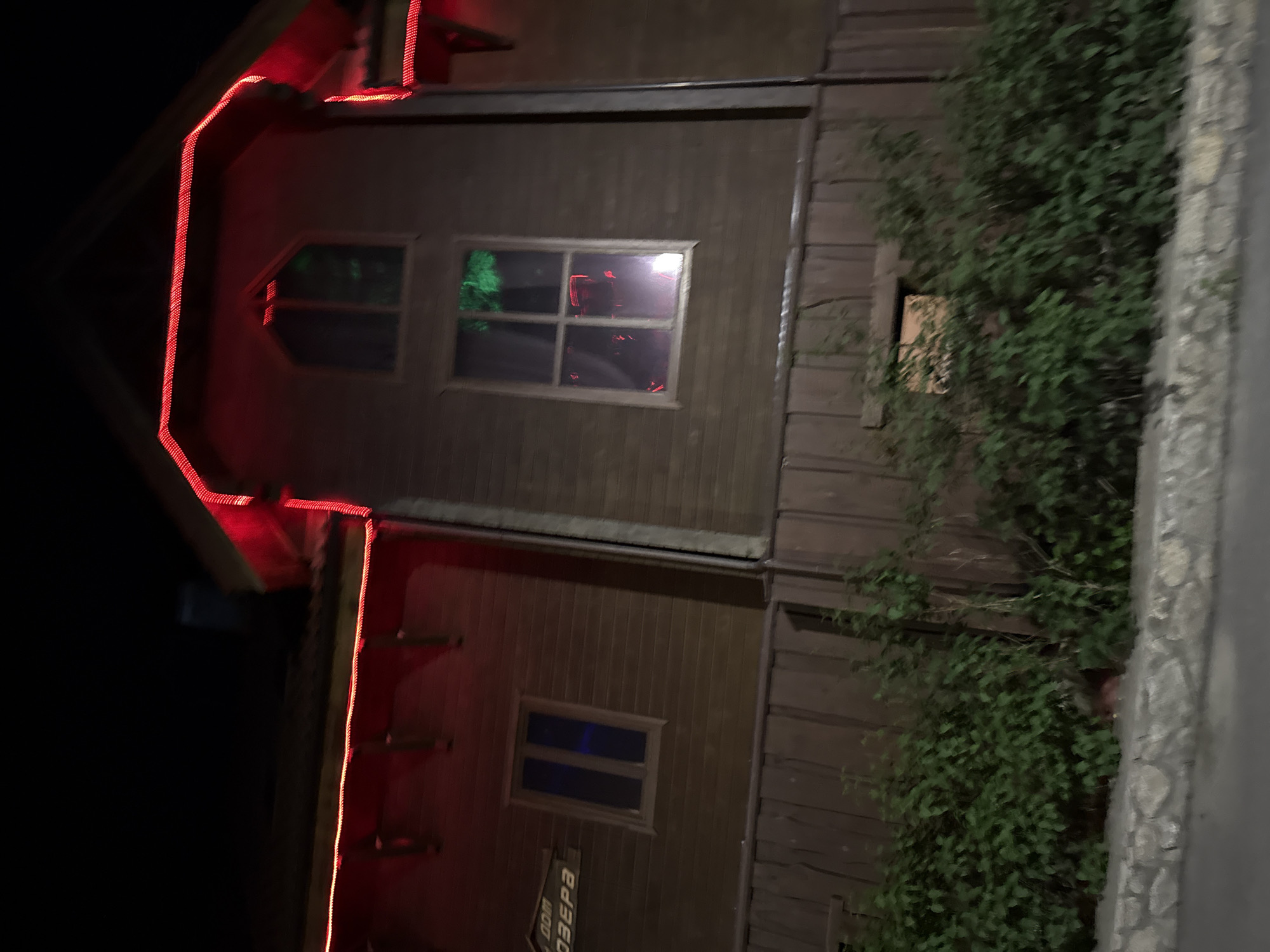 A nighttime exterior shot captures a wooden building adorned with red LED strip lights outlining its roofline and eaves. The building features several windows, some dark and reflective, others emitting a faint glow. One window, a multi-paned square, reflects green foliage and red lights from within. Above this window, a decorative arched window is visible. A sign with Cyrillic lettering, partially obscured, is mounted on the wooden facade. Lush green bushes and shrubbery grow in front of the lower portion of the building, partially obscuring a stone retaining wall. The overall scene suggests a building in an outdoor, possibly recreational, setting at night. The location is identified as Zolotoi Plyazh, Russia.