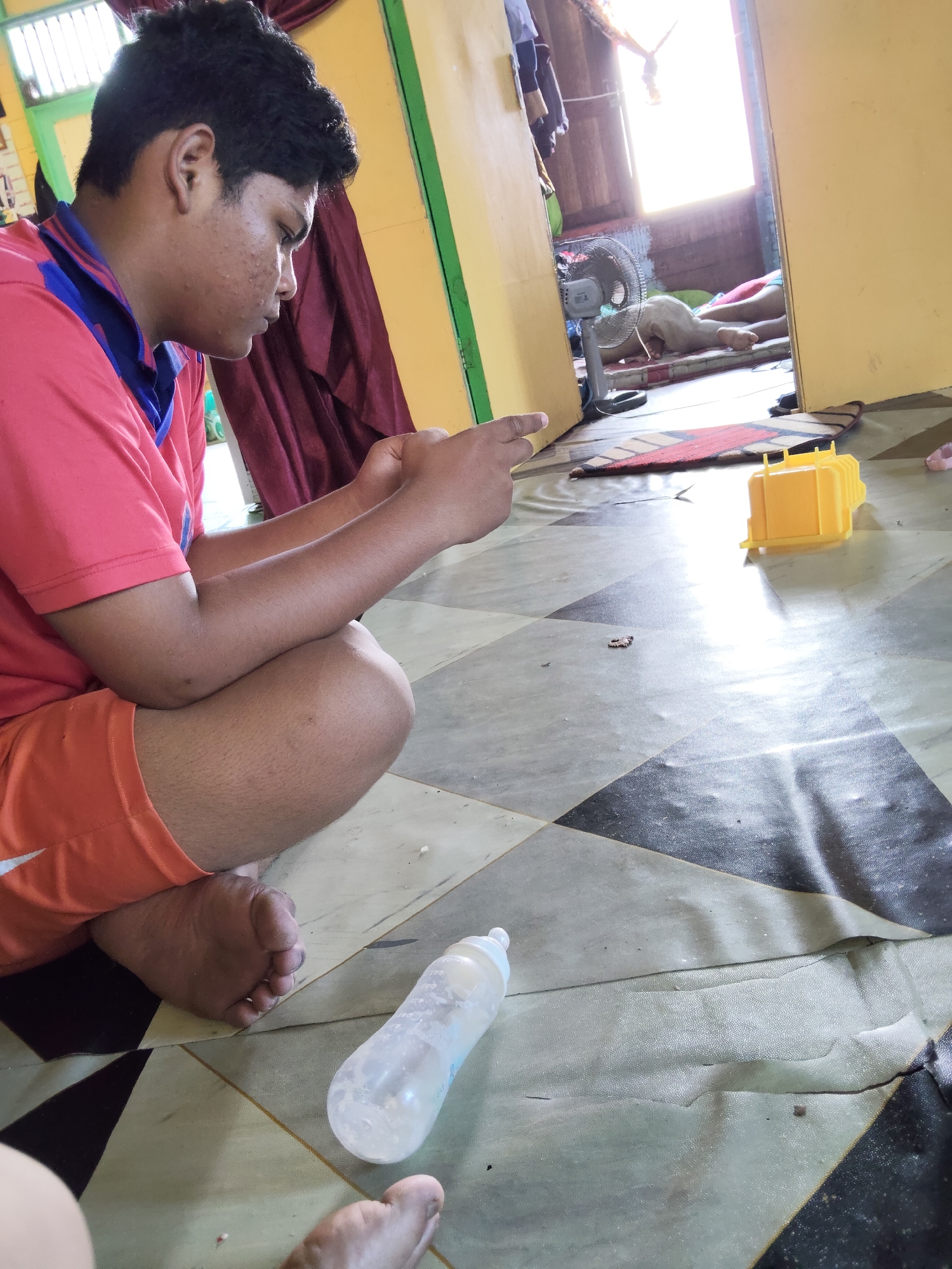 In an interior residential setting in Tanah Grogot, Indonesia, a young person is seated on a patterned floor, engrossed in a mobile device. They are wearing a coral-colored t-shirt with blue trim and orange shorts. Adjacent to the seated individual is a clear baby bottle. In the background, through an open doorway, another person is lying down on a mattress. A fan is positioned near the lying person, and sunlight streams in from a window. A yellow plastic toy is visible on the floor near the center of the frame. The floor is covered with a large, irregularly shaped mat with a checkered pattern of black and grey squares.