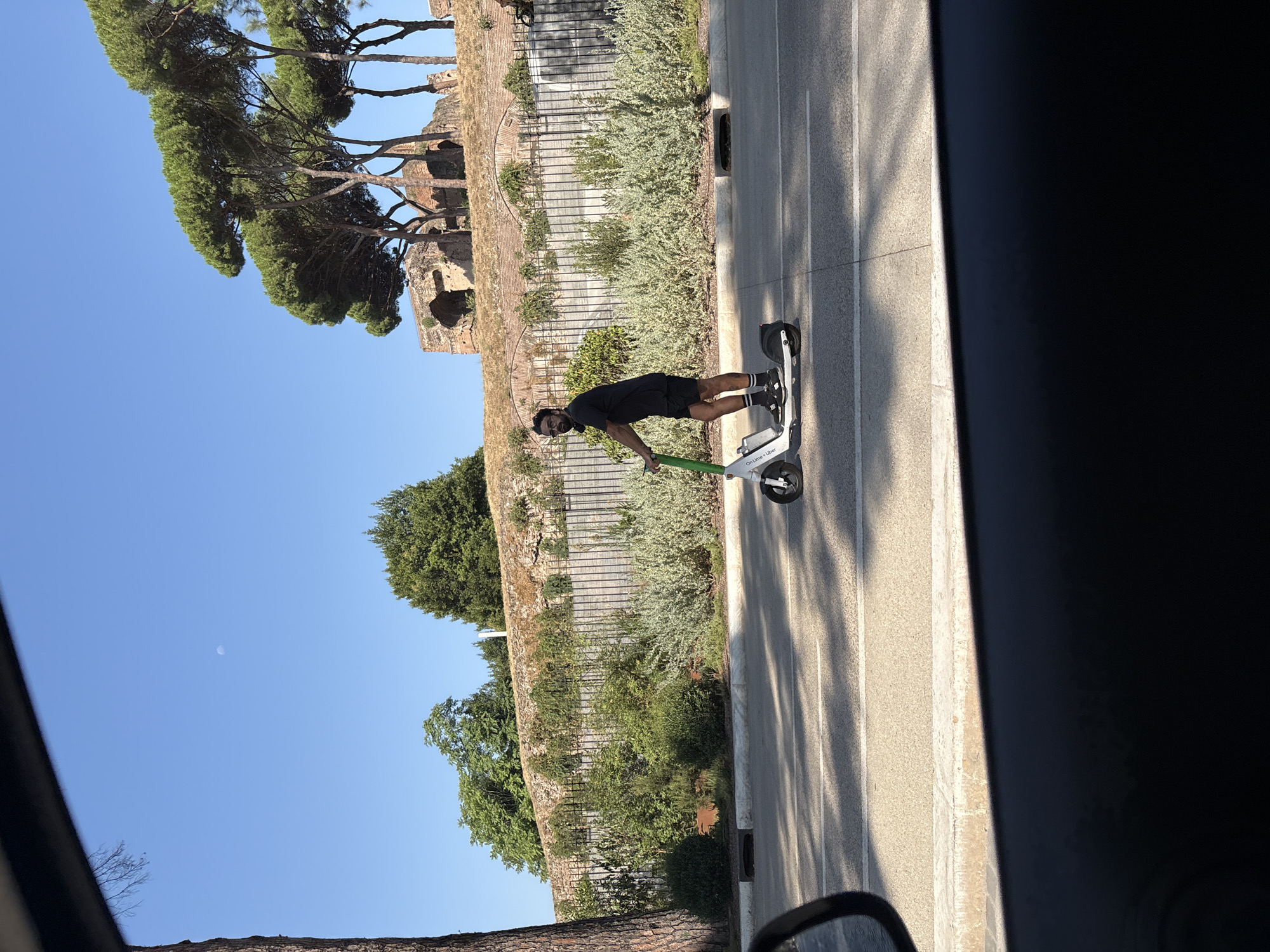 A man is riding a white electric scooter with green handlebars on a paved road in Rome, Italy. The scooter has "Lime" written on its side. The man is wearing a black t-shirt, black shorts, black and white socks, and black shoes. He also has black headphones around his neck and is wearing sunglasses. He is facing to his right. The road is lined with a curb and then a grassy area with dense green bushes. Behind the bushes is a black metal fence, and behind that is a stone wall with ancient Roman ruins on top, including brick arches. Tall pine trees are visible above the ruins. The sky is clear and blue with a faint moon visible. The image is framed by a dark interior, possibly a car, with a rearview mirror on the left side. Shadows of trees and possibly a vehicle are cast on the road.