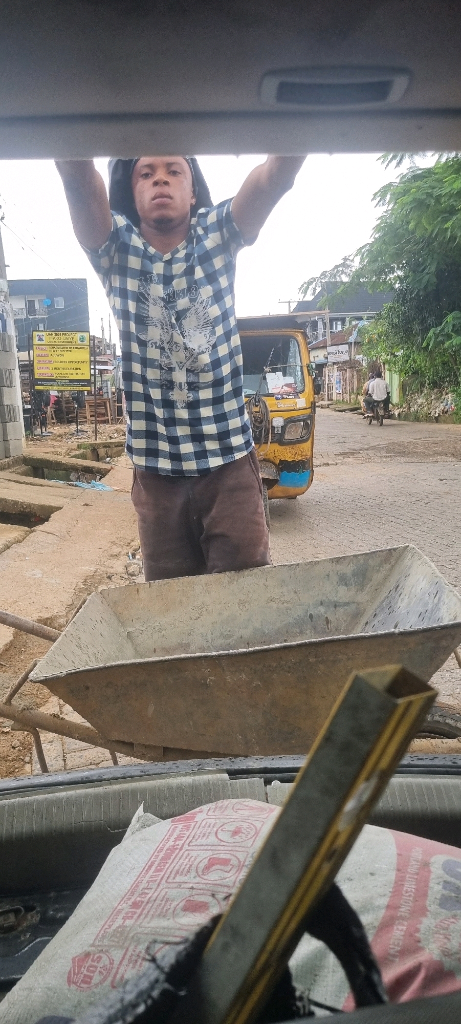 The image shows a young man standing with his hands on the top edge of an open car trunk. He is wearing a blue and white checkered shirt with a graphic print and brown shorts. In the background, a yellow auto-rickshaw is parked on a street. A construction sign is visible to the left of the auto-rickshaw, which reads "UIAH 2026 PROJECT IFAKO-IJAYE LOCAL GOVERNMENT". The street appears to be in a semi-urban area, with buildings and trees visible. A wheelbarrow is positioned in the foreground, within the car trunk. The overall scene suggests a location in Iju, Nigeria, likely a street with ongoing construction or development.