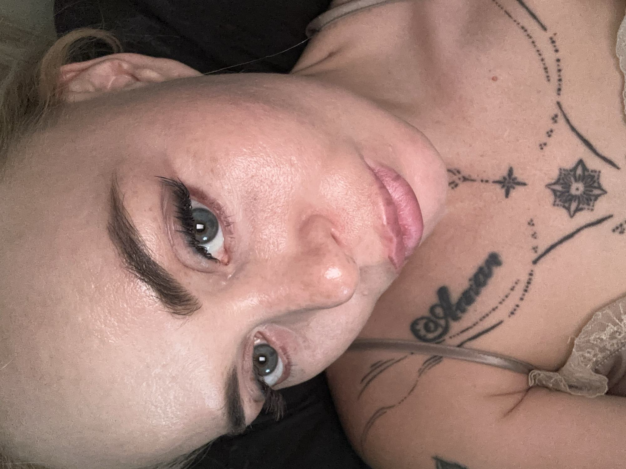 The image is a close-up, overhead shot of a woman's face and upper chest. She has light skin with some redness and texture. Her eyes are a greenish-blue color, and she is wearing false eyelashes and light makeup. Her eyebrows are dark and well-defined. Her lips are slightly parted and appear to be pink. She has blonde hair pulled back. There are several black tattoos visible on her chest and shoulder. One tattoo on her shoulder appears to be script that reads "Arman". Below her collarbone, there are geometric tattoos, including a star and a mandala-like design. She is wearing a light-colored lace bra that is partially visible. The background is dark and out of focus. The scene is indoors, and the lighting suggests it might be artificial light.