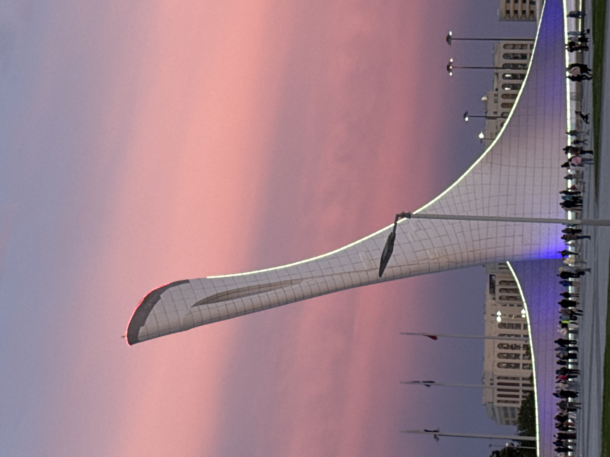 The image shows a large, white, arching structure, possibly part of a monument or building in the city of Sirius, Russia, with a grid-like pattern on its surface. The arch is illuminated along its edge with a blue light. A crowd of people is gathered along a walkway adjacent to the structure. Several flagpoles with flags are visible in the foreground, and a large building is partially visible in the background. The sky in the background is a mix of pink and purple hues, indicating either sunset or sunrise. The image is rotated.
