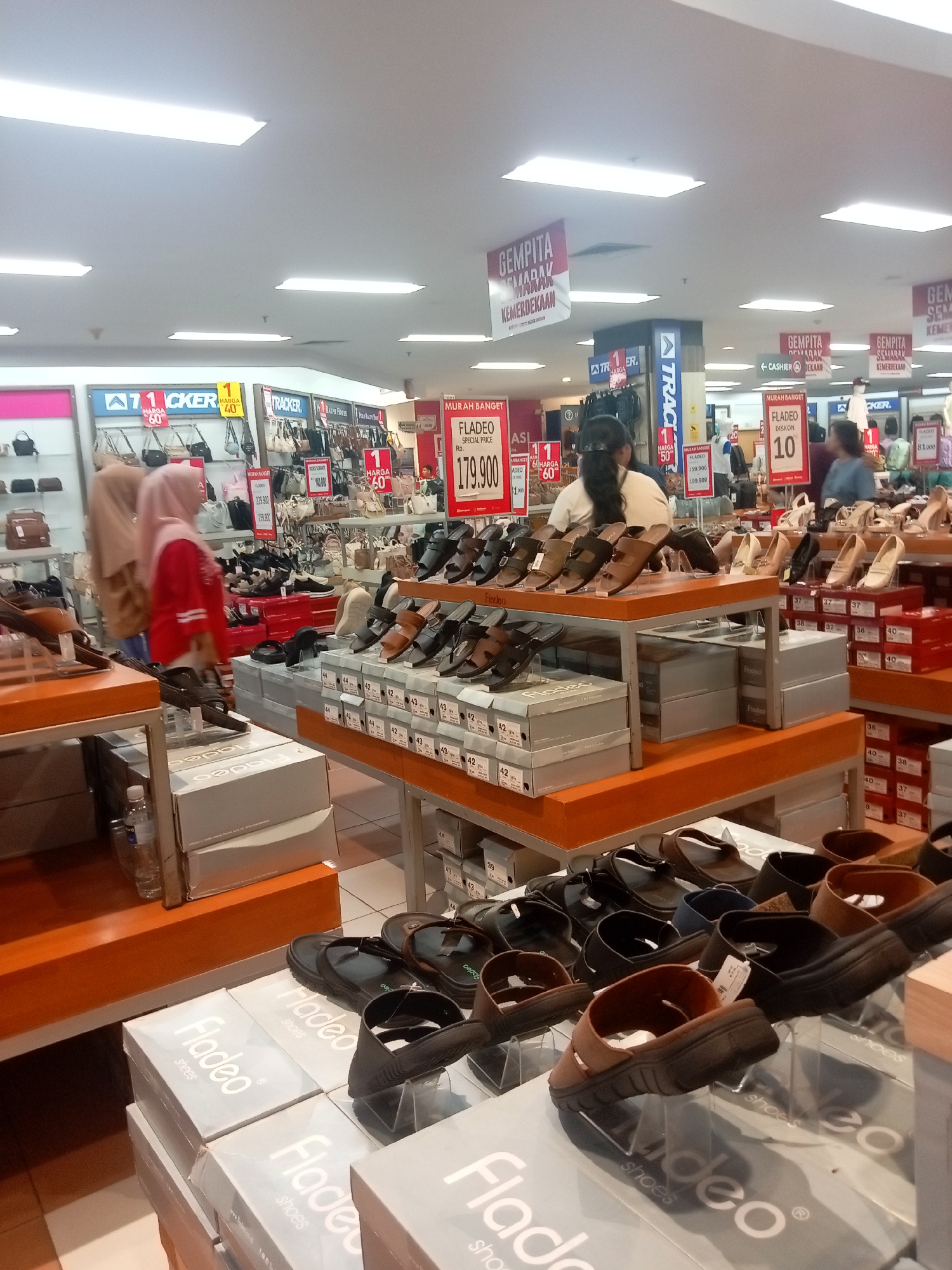 The image depicts an indoor shoe store, likely in Batam, Indonesia, given the visible Indonesian signage. The store features a wide selection of women's footwear displayed on shelves and tables. Several signs advertising discounts and promotions are visible, including "GEMPITA SEMARAK KEMERDEKAAN" (Festive Independence Day) and various price reductions. Two women, one wearing a hijab and a red top, and another wearing a light pink hijab, are browsing the merchandise. Another woman with dark hair is visible behind a counter. The store appears well-lit with overhead fluorescent lighting. Boxes with the brand name "Fladeo" are stacked in the foreground, suggesting a focus on this particular brand. The overall environment is that of a retail setting with organized displays of shoes.