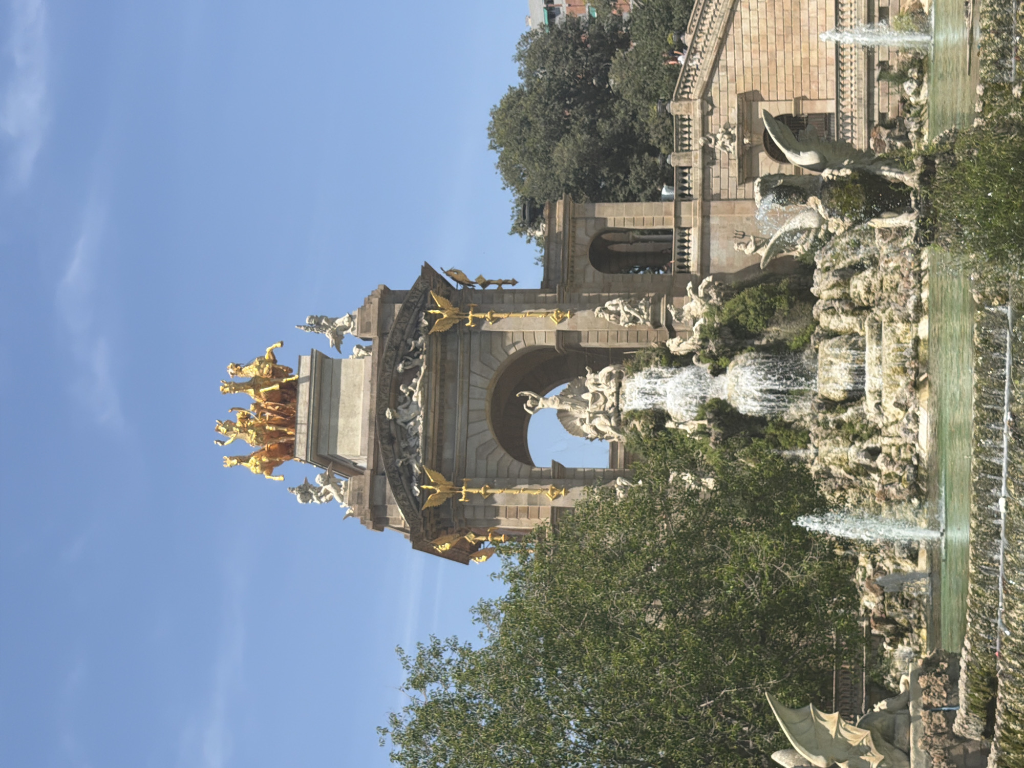 This is a wide-angle, eye-level shot of the Cascading Fountain in Parc de la Ciutadella, Barcelona, Spain, on a sunny day. The sky is a clear blue with a few wispy clouds.
The central focus is the monumental fountain, a complex Baroque structure adorned with golden statues and cascading water. At the very top, a golden chariot pulled by four horses is illuminated by the sun. Below this, a statue of a figure, possibly Neptune, holds a trident. The fountain itself is composed of multiple tiers of sculpted rock and water features, with water flowing down over the rocky surfaces.
To the left of the fountain, a large, leafy green tree dominates the frame, partially obscuring the structure. On the right side of the fountain, a grand stone staircase with balustrades leads up to a building. The staircase is lined with people walking up and down.
In the foreground, a pool of water reflects the fountain and the surrounding greenery. Several smaller fountains are active in the pool, sending streams of water into the air. A stone sculpture of a winged creature, possibly a dragon or griffin, is situated in the pool on the right side of the frame.
The architecture of the fountain is intricate, with arched openings, sculpted figures, and decorative elements in stone and gold. The overall scene conveys a sense of grandeur and natural beauty, typical of a public park in a European city.