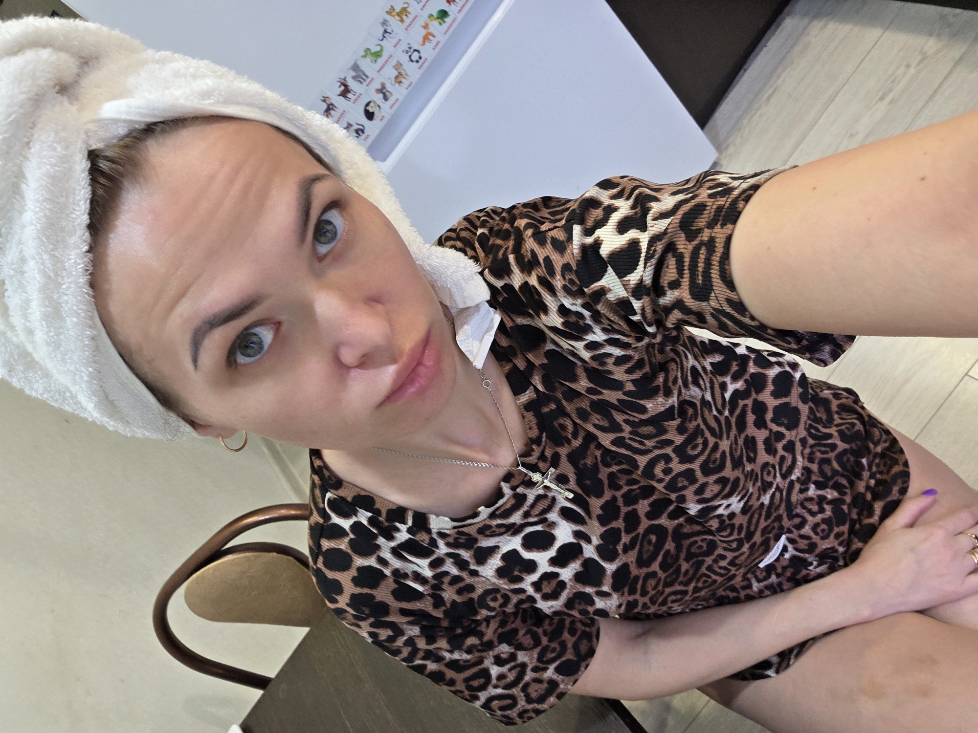 A selfie depicts a woman with a white towel wrapped around her head. She has fair skin, light eyes, and visible eyebrows. The woman wears a leopard print top and a silver necklace with a cross pendant. She is seated on a chair, visible in the lower left of the frame, with a wooden back and tan seat. She is wearing two rings on her fingers with purple nail polish. Behind her is a white surface with various animal stickers near the top of the frame. The location is possibly in a residential setting, and the city of Chaikovsky, Russia, is unknown.
