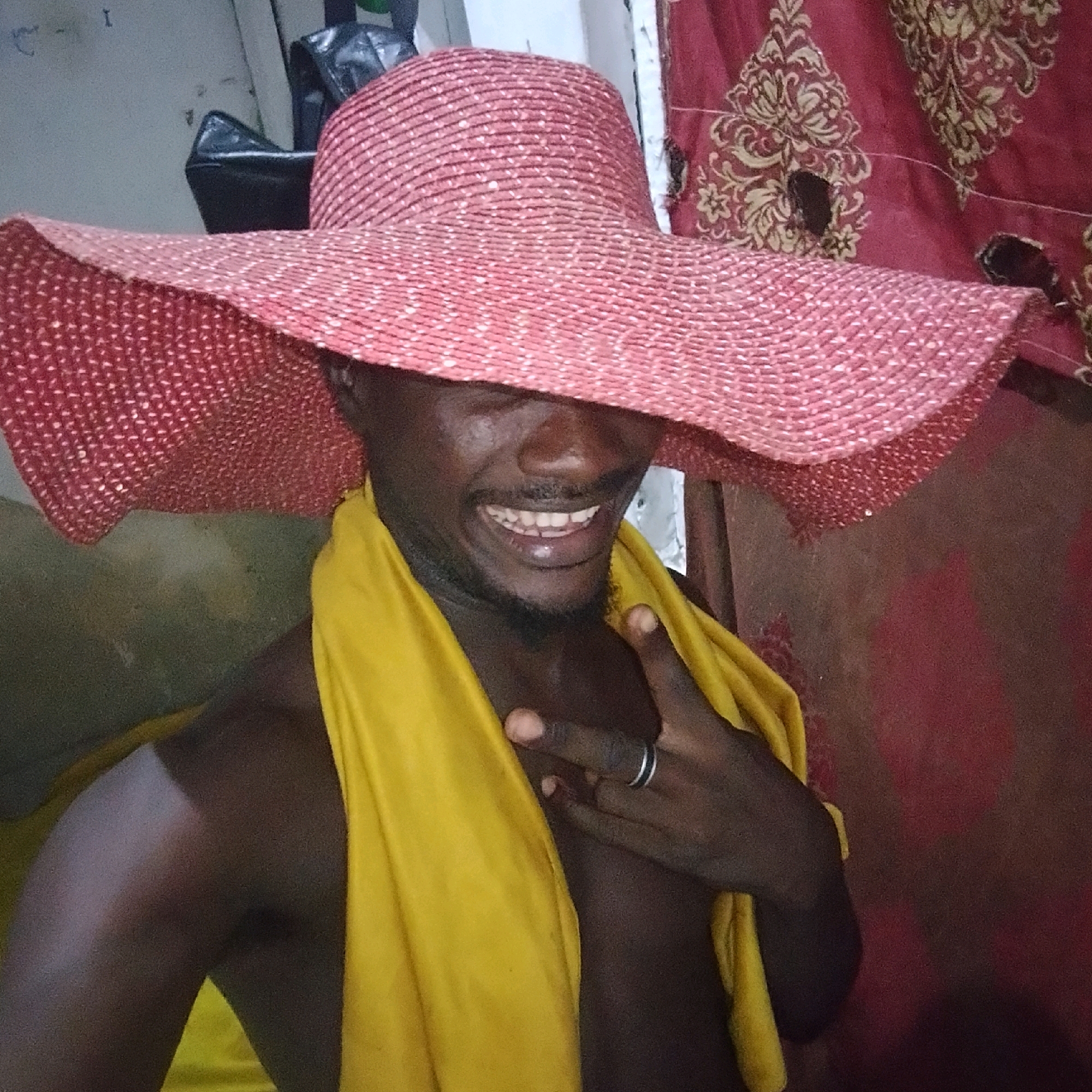 A person, identified as being in Ado, Nigeria, is captured in a medium shot, wearing a large, red, wide-brimmed hat with a textured pattern. They are draped in a bright yellow fabric that covers their shoulders and chest, with one hand visible, adorned with a ring on one finger and positioned near their chest. The background is a dark, patterned fabric, possibly a curtain or tapestry, with holes or tears visible in it. The lighting appears to be dim, creating shadows on the person's face and body. The overall scene suggests an indoor environment, possibly a room.