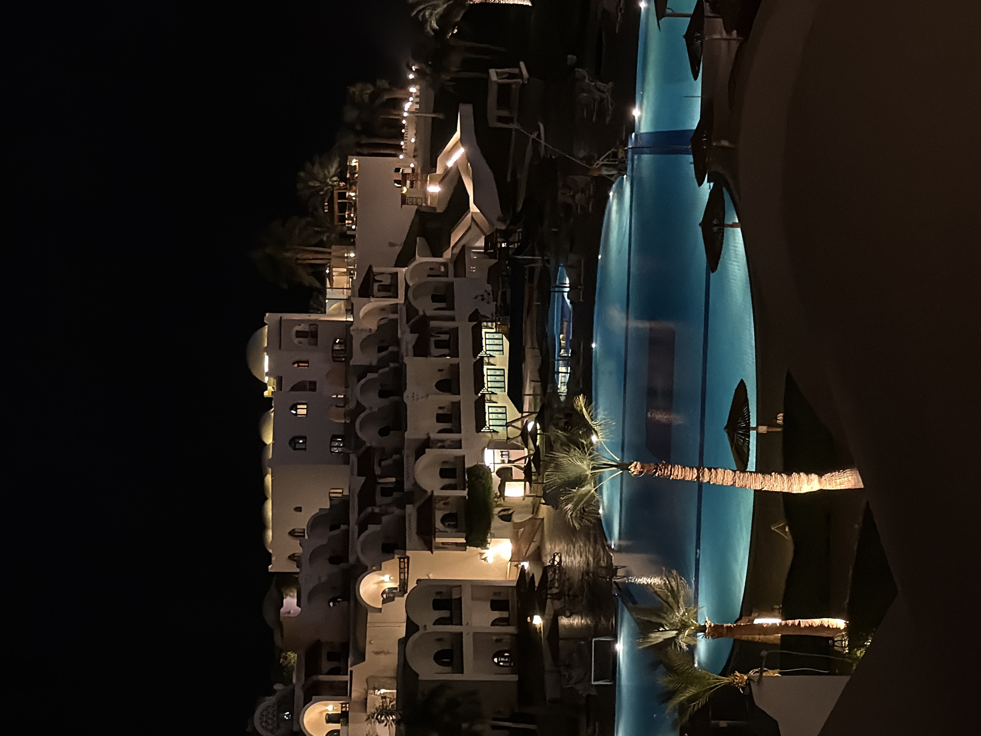 Nighttime view of a resort in Sharm El Sheikh, Egypt, with a large swimming pool in the foreground. The pool is illuminated and reflects the lights from the surrounding buildings. The buildings are multi-story with arched windows and balconies. Palm trees are visible along the edge of the pool and throughout the resort. There are no people visible in the scene.