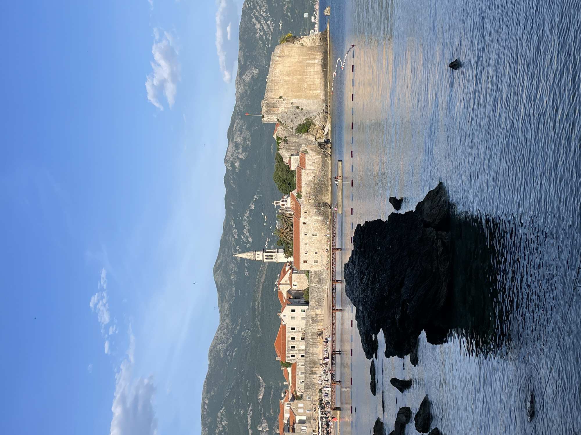 The image shows a side view of the Old Town of Budva, Montenegro, with the Adriatic Sea in the foreground. The old city is constructed on a peninsula with stone walls and buildings. A tall, white bell tower is visible, part of the Church of St. Ivan. Mountains rise behind the city. Rocks jut from the sea, and a few people are visible along the edge of the city. The sky is blue with some clouds.