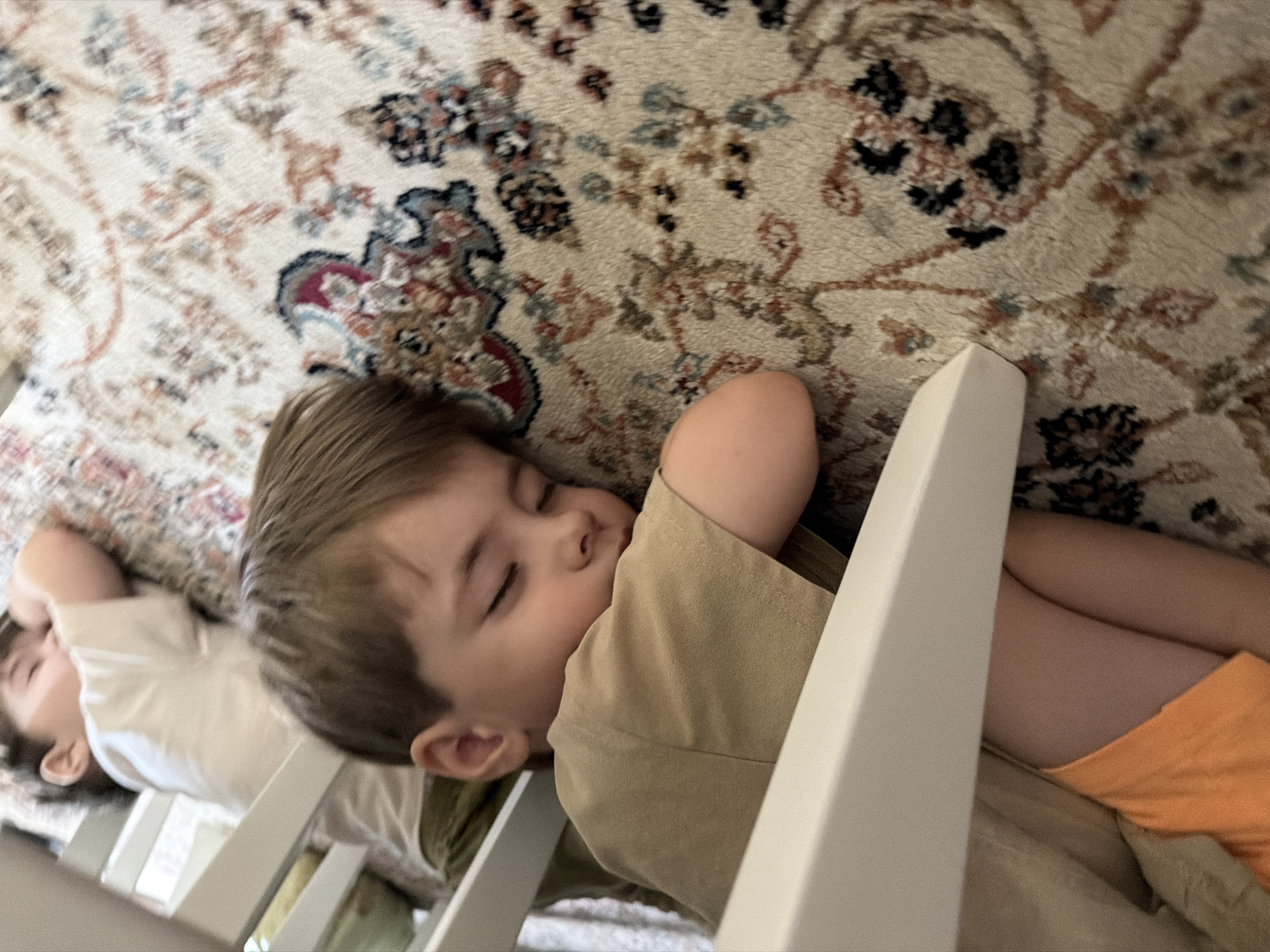 Two young children are sleeping on a carpeted floor. The child in the foreground is lying on their side, with their head resting on their arm, wearing an orange pair of shorts and a tan t-shirt. Their eyes are closed. The second child is lying in the background on their stomach, also with their eyes closed, wearing a white t-shirt. Both children appear to be under a white piece of furniture, possibly a table or bed frame. The carpet has a floral pattern with cream, red, blue, and black colors. There is no information in the image to suggest this scene is located in Malgobek, Russia.